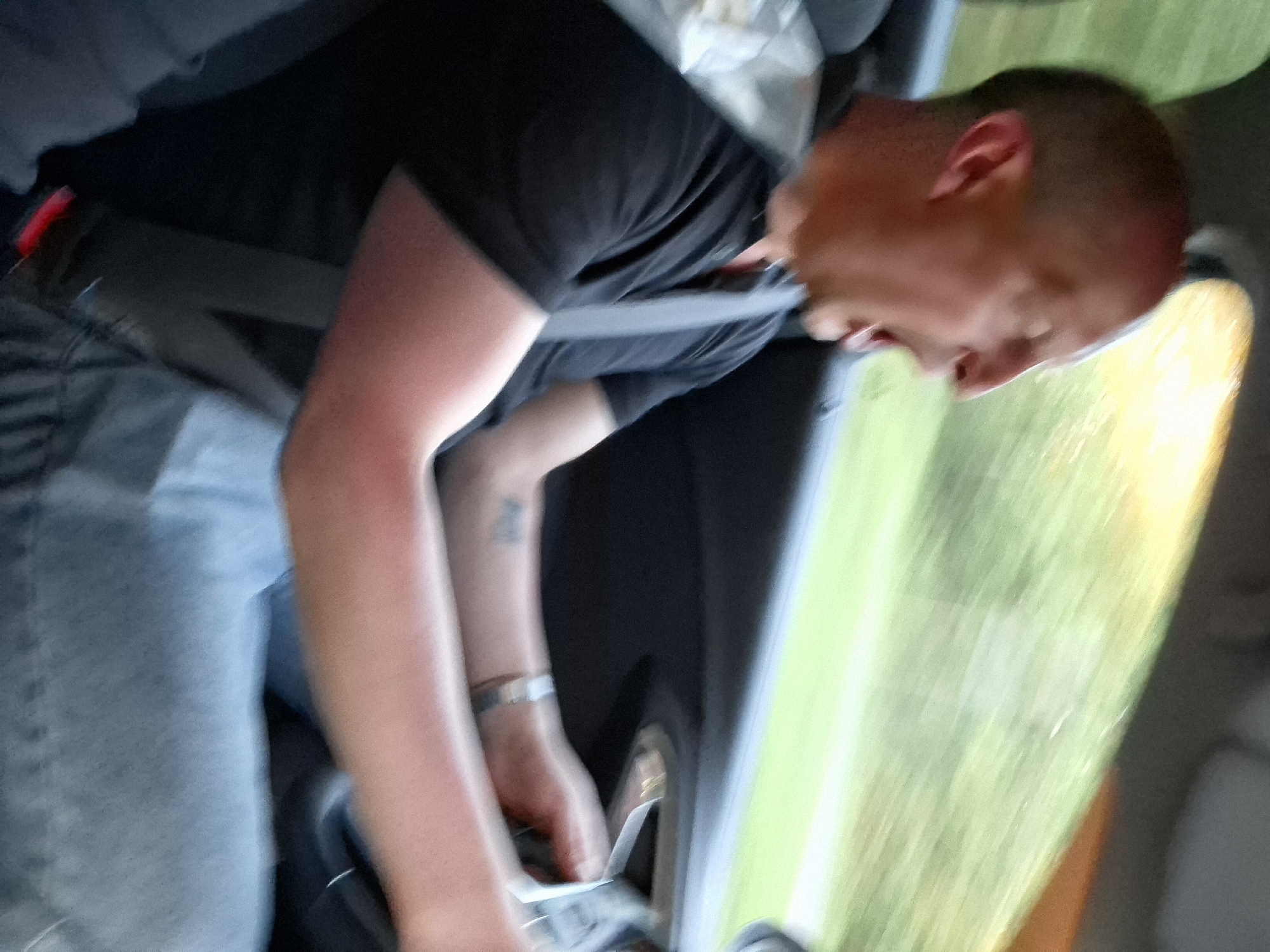A man wearing a black shirt and blue jeans is seated in a car, secured with a seatbelt. The vehicle appears to be in motion, indicated by the blurred view of green and yellow landscape visible through the car window. The man is likely reaching for the car door handle. The image does not provide enough detail to confirm the exact location in Любанскі сельскі Савет, Belarus.