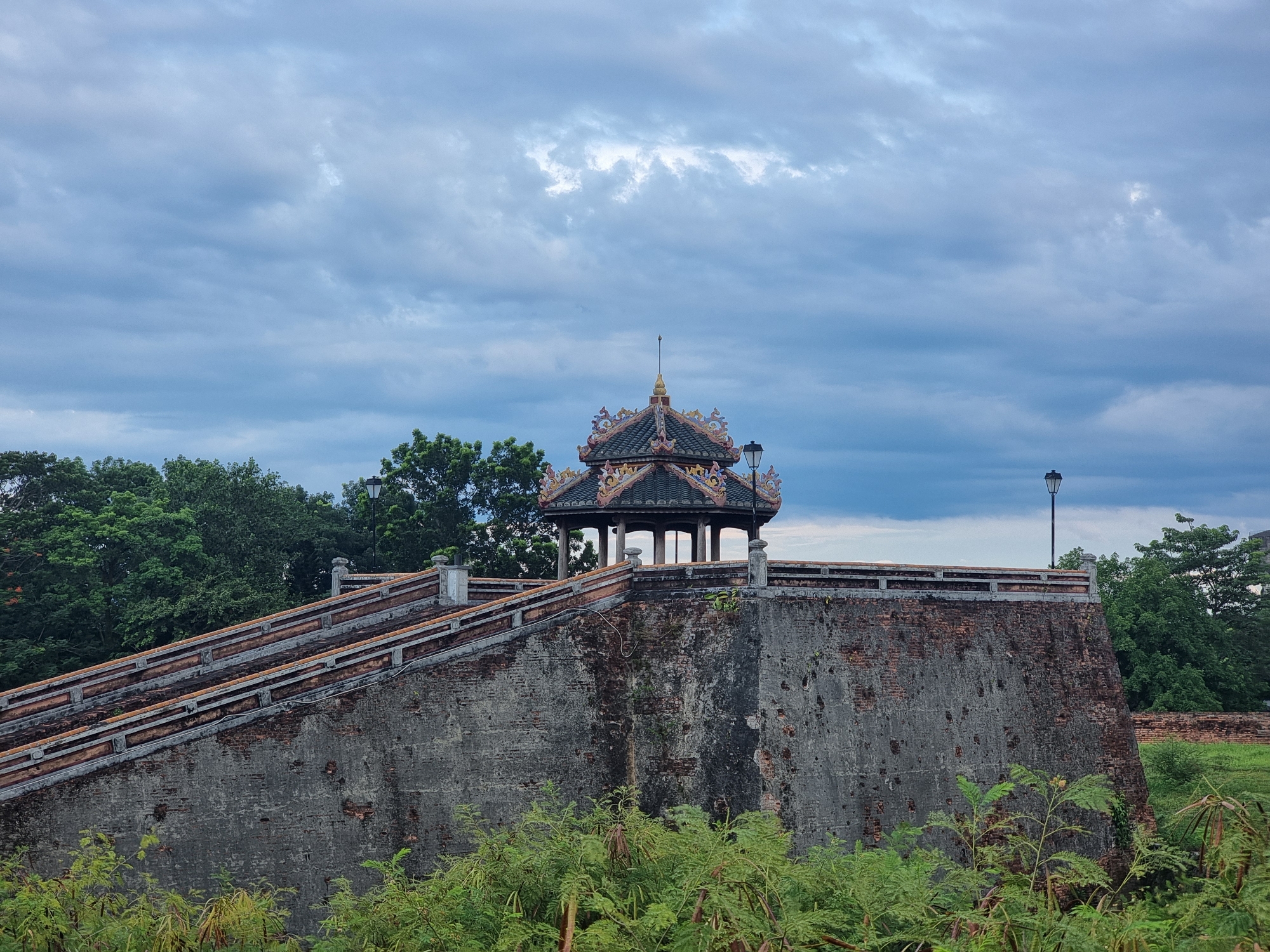 A weathered, grey brick fortress wall dominates the lower half of the frame, its surface showing signs of age and damage, with patches of exposed red brick and dark stains. A sloping, tiled ramp with stone railings ascends from the left side of the wall. Atop the wall, an ornate, multi-tiered pavilion with a dark, tiled roof, adorned with decorative motifs and a spire, is visible. A lamppost stands on the right side of the wall. Lush green foliage, including bushes and trees, frames the lower and upper portions of the scene. The sky above is overcast with heavy, grey clouds. The location is the Citadel in Phú Xuân Ward, Vietnam.