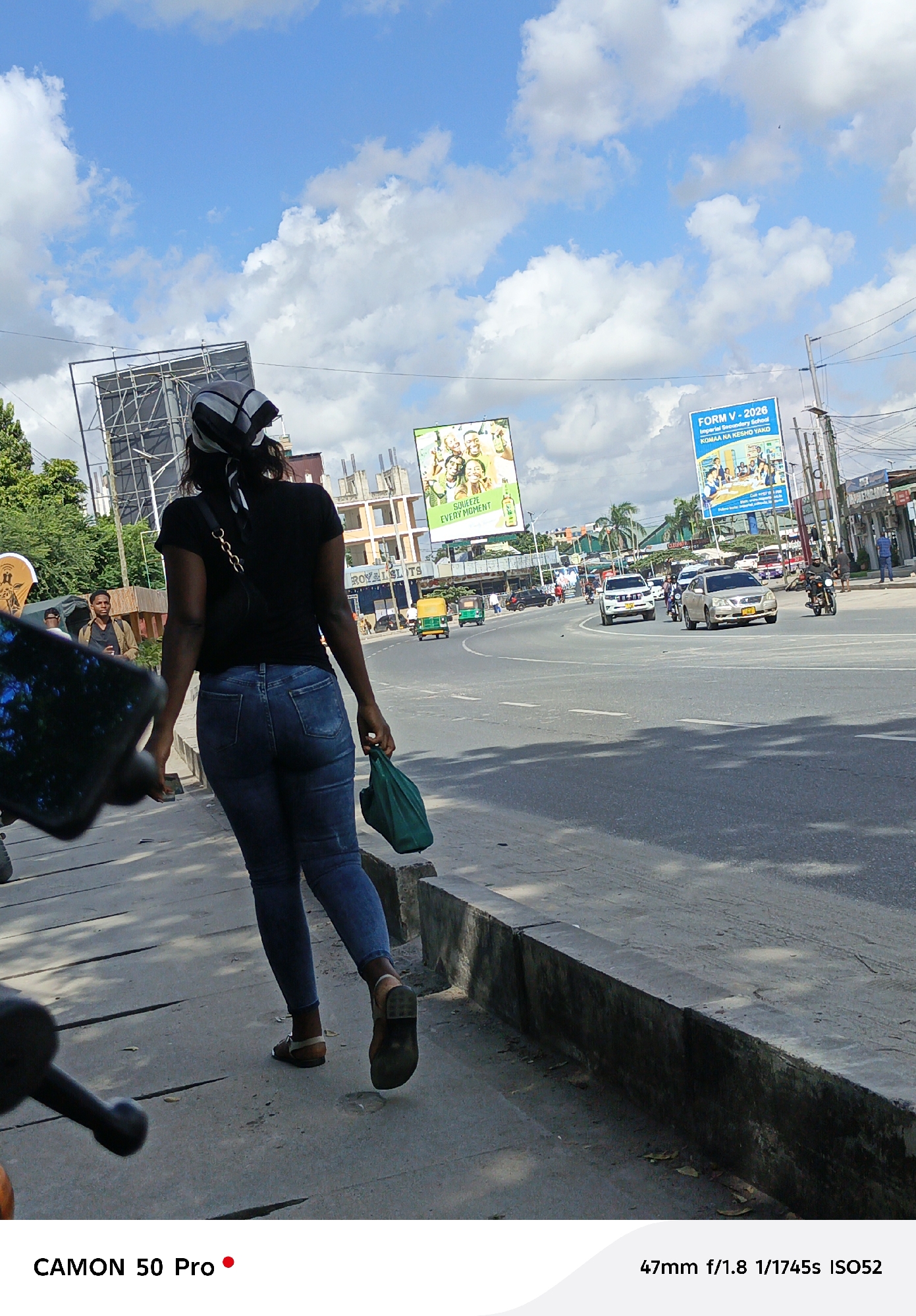 The image depicts a street scene in Dar es Salaam, Tanzania, featuring a woman walking on a sidewalk alongside a busy road. The woman, seen from behind, is wearing a black t-shirt, blue jeans, sandals, and a headscarf. She is carrying a green bag. On the road, there are several vehicles, including cars, auto-rickshaws (tuk-tuks), and motorcycles. Billboards line the street, one advertising a beverage ("SQUEEZE EVERY MOMENT") and another promoting a secondary school ("FORM V - 2026"). There is a building with visible construction in the background. The sky is blue with scattered clouds. A partial view of a camera or smartphone is visible in the foreground, indicating the image was taken with a mobile device.