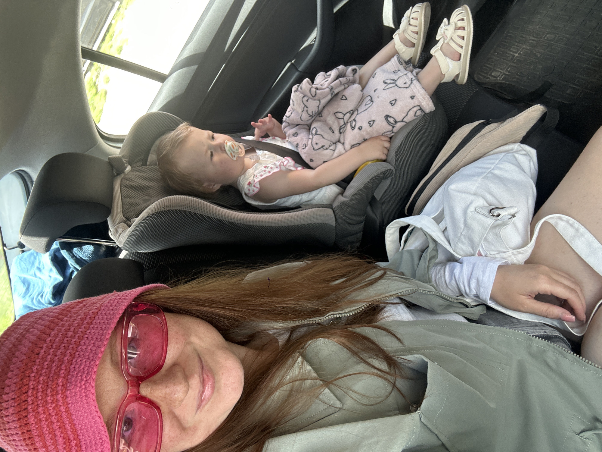This image captures a relaxed scene inside a car, likely in Nizhny Novgorod, Russia, during daytime. The perspective suggests a selfie taken by a woman, whose face and upper body are partially visible in the foreground on the left. She wears a vibrant pink knitted hat and red-framed sunglasses, with long, reddish-brown hair cascading over her shoulders. She is dressed in a light green quilted jacket over a white long-sleeved top, and her expression, though partially obscured, appears calm.
In the background, a young child, likely a toddler, is nestled comfortably in a gray and black car seat. The child has light-colored hair and is wearing a light-patterned dress, with a pacifier in their mouth, suggesting they are resting or possibly sleeping. A soft, light pink blanket adorned with bunny and heart patterns covers their legs, and they wear light-colored sandals with small bows.
The car interior features dark upholstery. Outside the car window, bright sunlight illuminates lush green foliage, indicating clear weather during the day. A beige fanny pack or small bag is visible on the car floor near the child's feet, and a blue fabric item can be seen on another seat further back. The overall impression is one of a peaceful car journey. No visible text is present in the image.