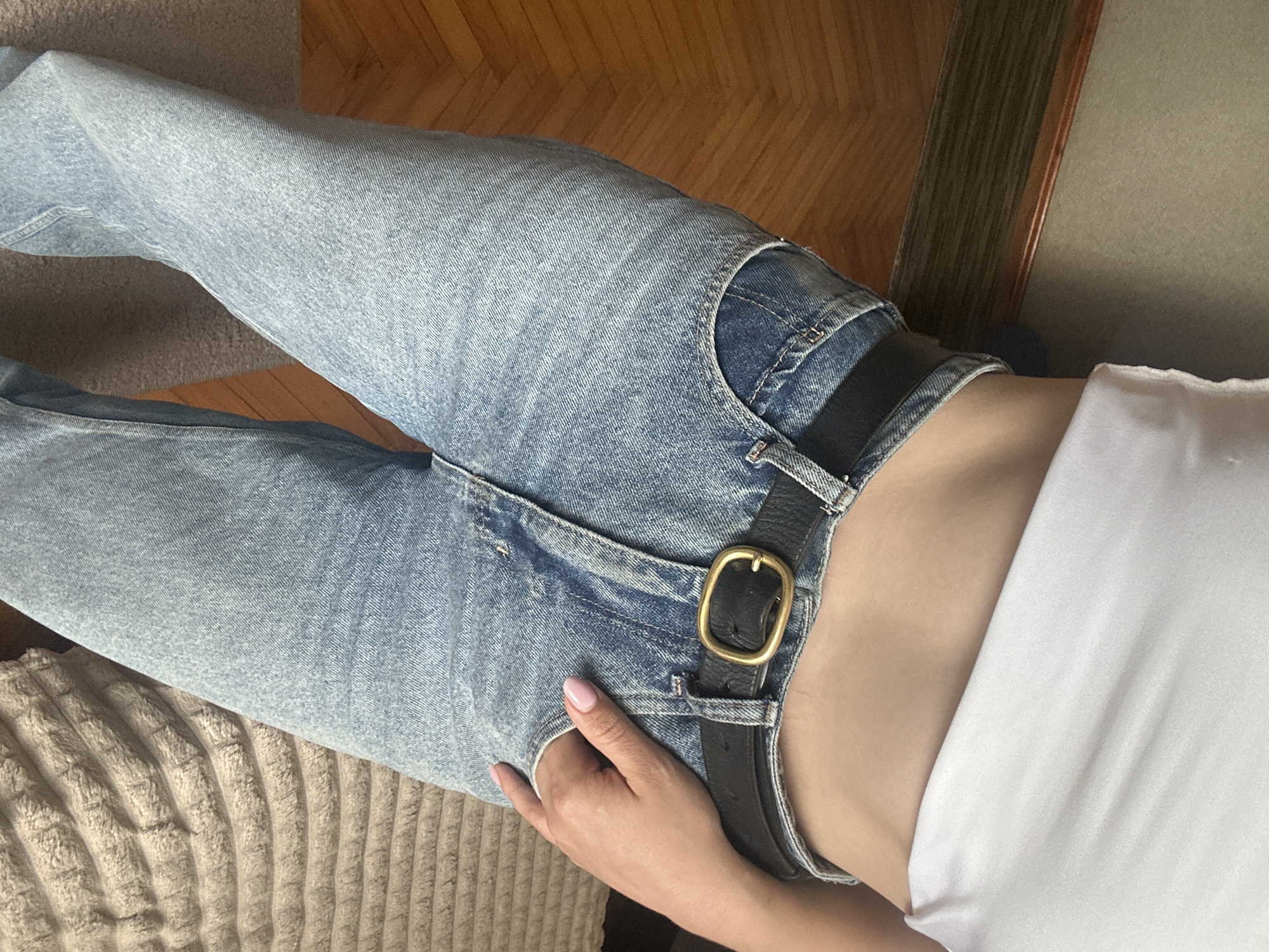 This close-up, high-angle shot captures the lower torso and legs of a person, likely reclining or lying down indoors in Ternopil, Ukraine. The primary subject is wearing light wash blue denim jeans with a black leather belt featuring a prominent gold or brass oval buckle. A simple white top is visible, extending from the bottom of the frame and slightly exposing a small section of the person's midriff near the belt. The person's left hand, with light pink manicured nails, rests on their right hip, with fingers subtly tucked into the jean pocket opening.
In the background, a light-colored, textured fabric, possibly a sofa or blanket, occupies the lower left portion of the image. The upper right reveals a wooden floor with a discernible grain pattern, suggesting parquet or floorboards. Natural, bright lighting suggests it is daytime. No specific activities or events are discernible beyond posing, and no text is visible in the image.