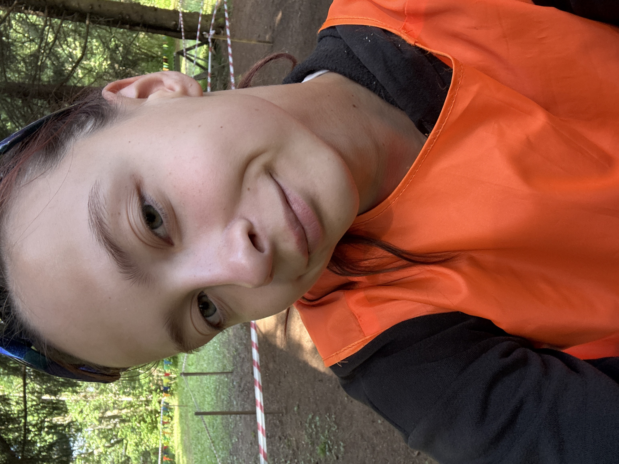 A young person, likely female, is captured in a close-up, low-angle selfie. The subject has light skin with freckles and is wearing an orange high-visibility vest over a black hooded garment. Their head is tilted, and they are looking towards the camera with a slight smile. Visible features include the left eye, a portion of the nose, lips, and ear. The background suggests an outdoor setting with trees and foliage, hinting at a wooded area or park. A red and white striped tape is visible in the lower part of the frame, possibly indicating a marked path or boundary. The lighting suggests it's daytime, with natural light illuminating the scene. The overall mood appears casual and perhaps adventurous, given the vest and outdoor environment. The location is identified as Sergino, Russia, which aligns with the natural, forested surroundings. No visible text is present in the image.