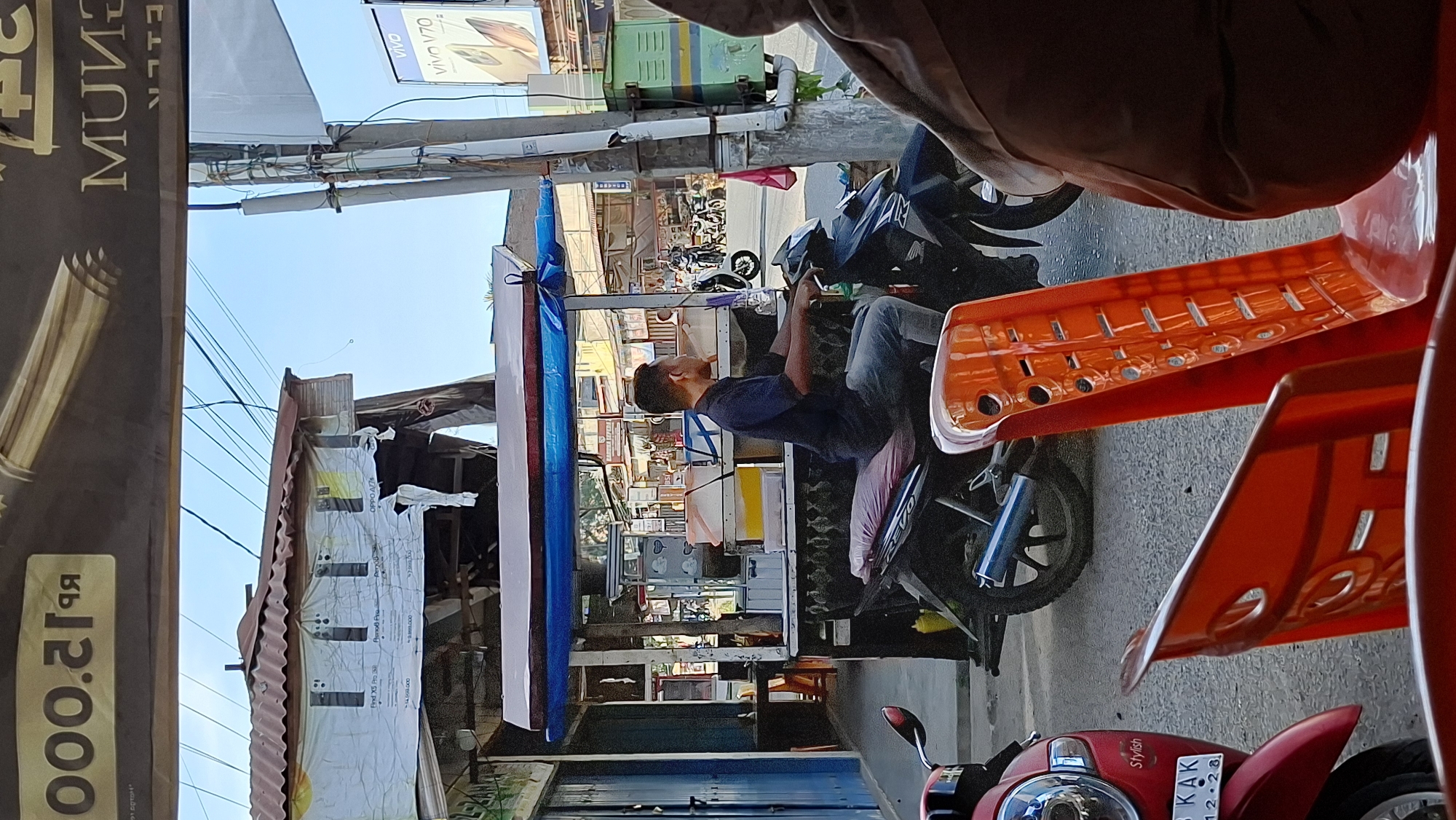 The image depicts a street scene in Keudebayu, Indonesia, likely in the morning or afternoon given the bright sunlight. In the foreground, a person is seated on a dark-colored motorcycle, wearing a dark blue t-shirt and blue jeans. The motorcycle is parked on a paved road, and a red scooter is partially visible to the left, with the license plate reading "8 KAK 12.28". Several orange plastic chairs are in the foreground, one of which is prominently displayed. Behind the motorcycle is a food stall with a blue and white awning, selling various items that appear to be food or drinks in containers. The stall has a metal roof and a wooden structure. In the background, there are other buildings and shops, suggesting a commercial area. A prominent banner across the top of the image displays "000.2199" and other text that is partially obscured or difficult to read. Another banner in the background advertises "vivo V70" and shows images of smartphones. The overall impression is a busy, everyday scene in a town or city.