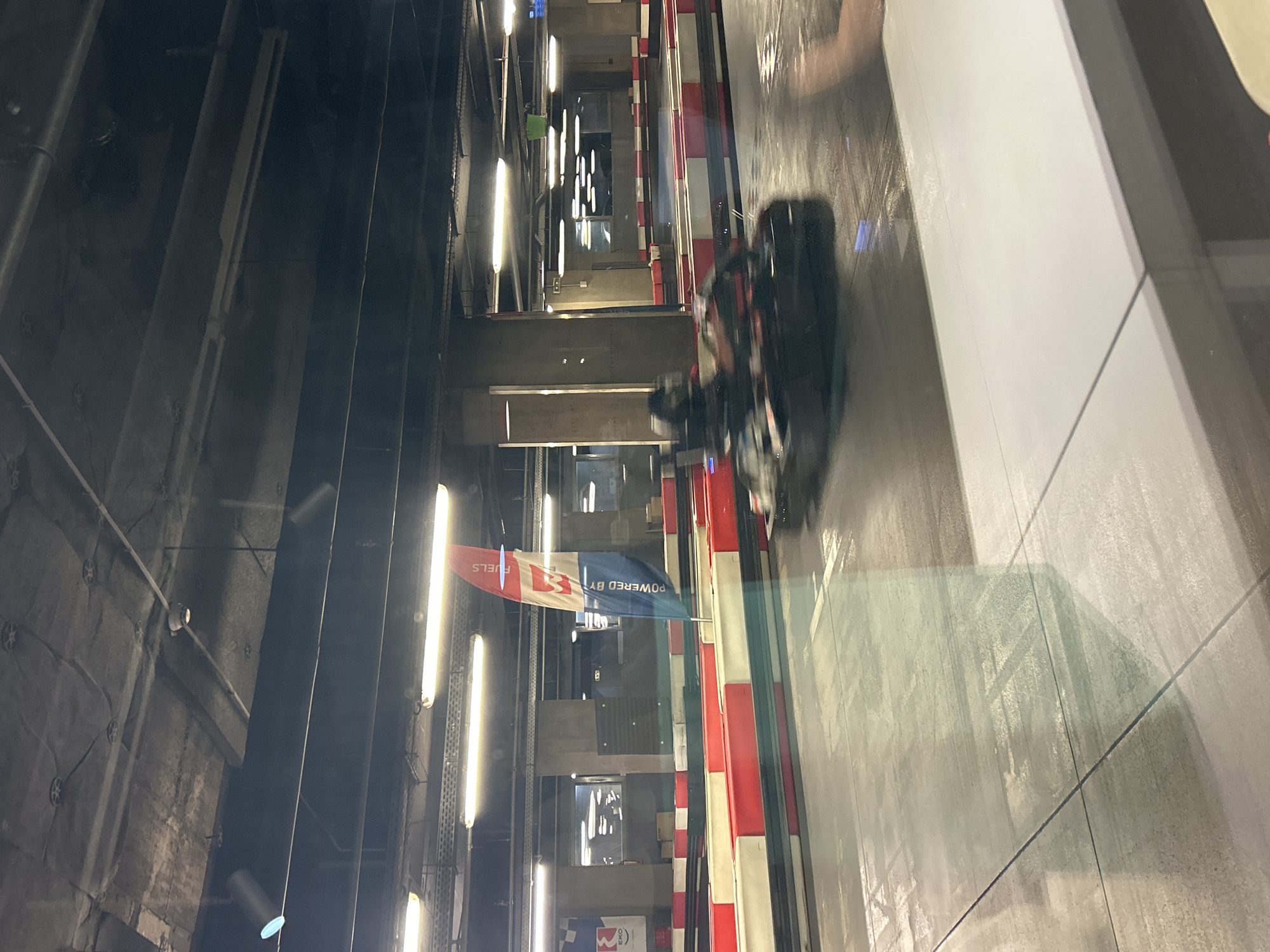 The image depicts an indoor go-kart track. The track is lined with red and white barriers. A go-kart, blurred due to motion, is speeding along the track. There are overhead lights illuminating the scene. A "Powered by EKO Fuels" flag is visible. The location is Karting Ring Sofia Ring Mall, Sofia, Bulgaria.