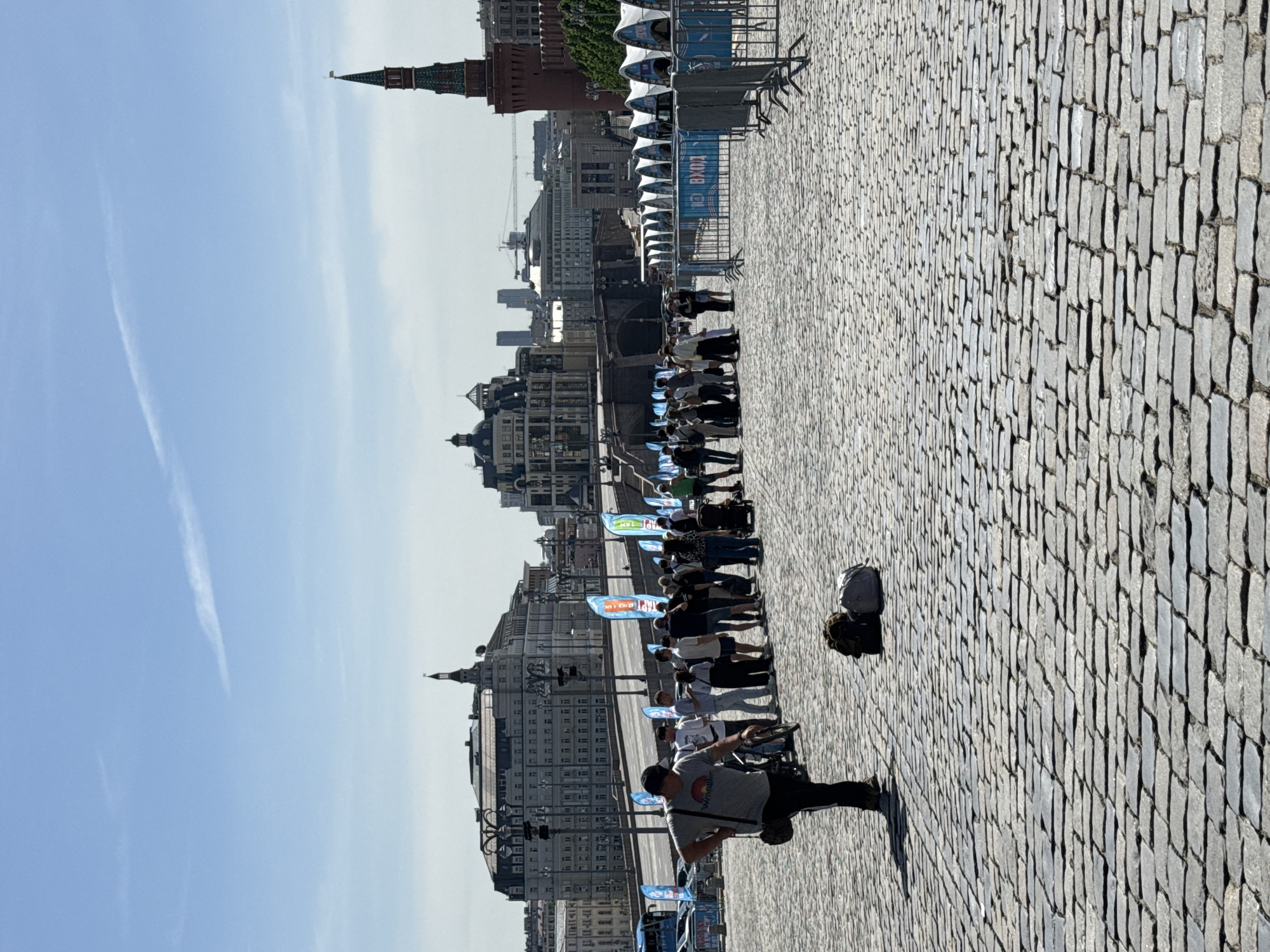 Here's a detailed description of the image:
**Scene Overview:**
The image captures an outdoor scene on a cobblestone plaza. The sky is clear and blue. A line of people is visible, possibly queueing or participating in an event. Buildings form a backdrop to the people.
**Location and Environment:**
The buildings in the background are characteristic of a European city, possibly in Russia. A red brick tower with a distinctive spire is visible, suggesting the presence of a historic structure, possibly the Kremlin.
**People:**
* A line of people stretches across the scene. Their faces are not clear.
* A person in grey is in the foreground.
**Objects:**
* Blue and white event flags.
* Barriers or railings.
**Actions and Interactions:**
* People are standing in a line.
**Overall Impression:**
The image suggests an event or organized activity taking place in a historic urban environment. The presence of barriers and flags indicates a structured event.