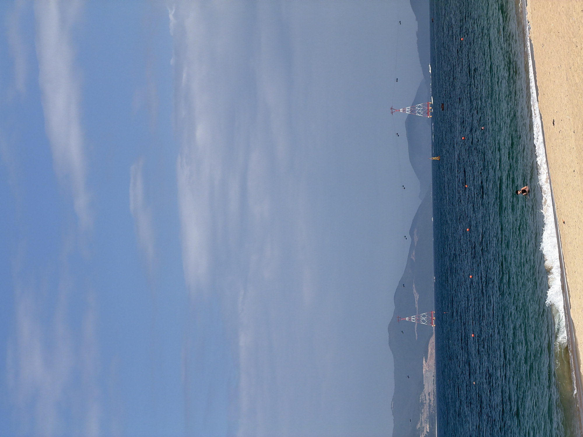 This image depicts a daytime coastal scene in Nha Trang Ward, Vietnam, observed from a sandy beach looking out towards the ocean and distant mountains. The upper portion of the frame shows a light blue sky with scattered white clouds. A range of hazy, dark green mountains forms the background across the horizon. The midground features deep blue-green ocean water with small white-capped waves breaking near the shore. Two prominent red and white lattice pylons, part of a cable car system, stand in the water in the middle distance, with multiple dark-colored cable car cabins visible traversing the overhead cables. Several orange spherical buoys and one yellow triangular buoy are dispersed across the water's surface, and a few small boats are visible further out at sea. In the immediate foreground, a strip of light brown sand defines the beach. Two individuals are present in the ocean near the shore; one person, wearing a dark top, is partially submerged on the right side, and another individual is partially submerged further out in the water.