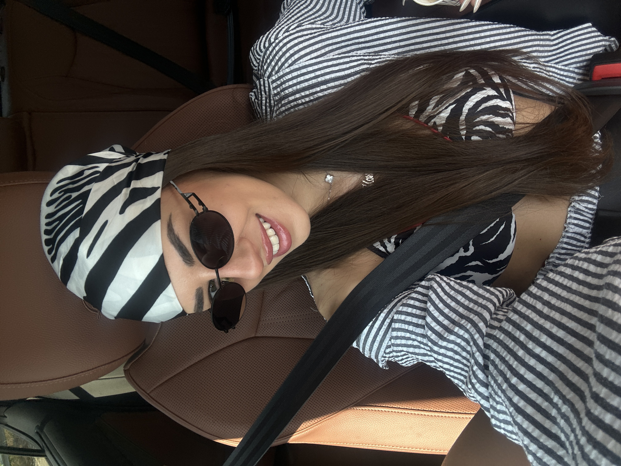 A woman with long, dark hair and a zebra-print headscarf is pictured inside a car, wearing sunglasses and a striped shirt. She is smiling at the camera, and her seatbelt is fastened. The interior of the car appears to be made of brown leather. The background shows car seats and a seatbelt. The image does not contain any information that can identify the location as Chryso, Greece.