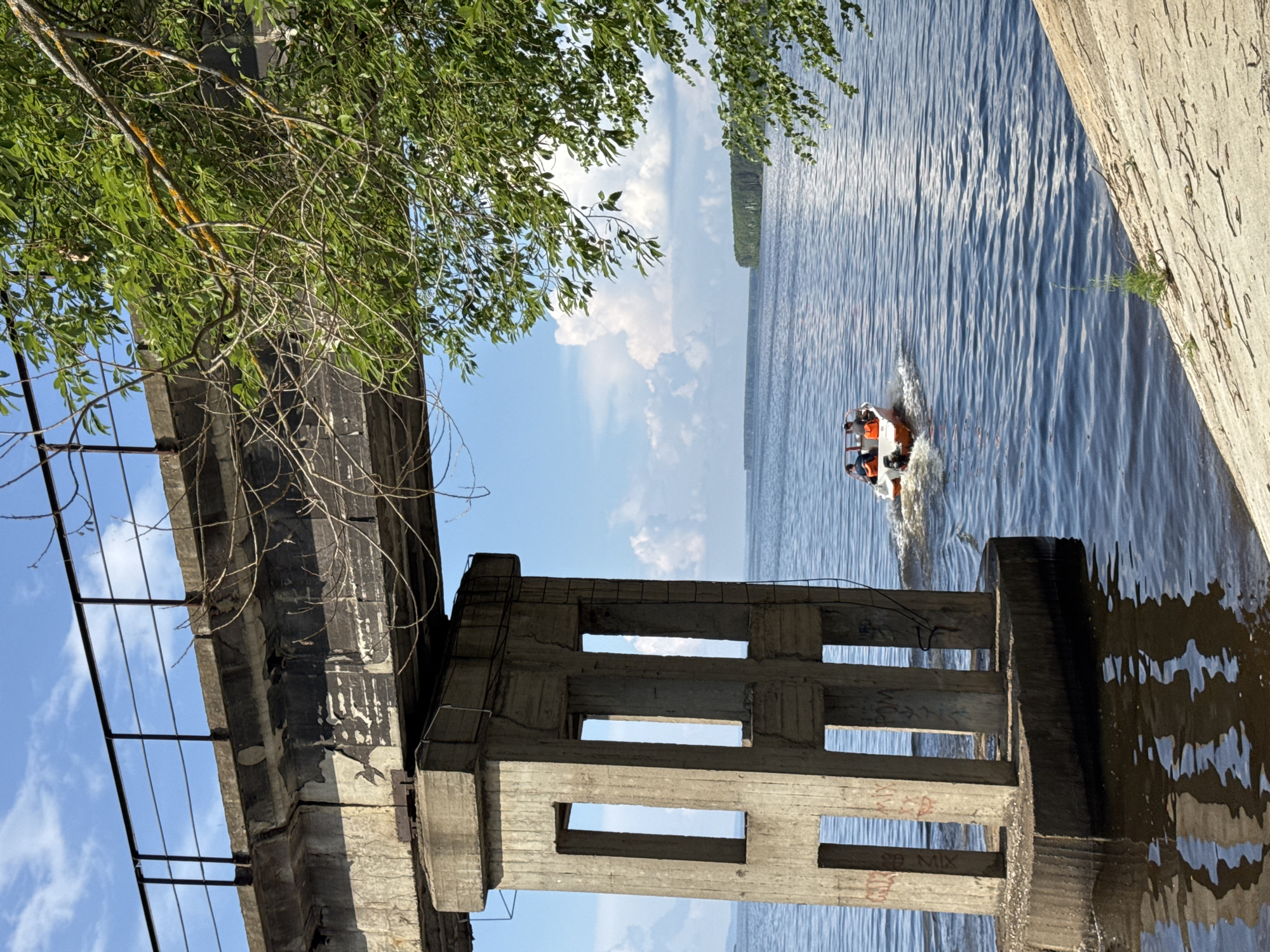 A concrete bridge pier with a railing and weathered concrete beam stands in the foreground on the left, casting a reflection on the dark blue water. In the middle ground, a small, orange and white motorboat with two people wearing life jackets travels away from the viewer on a wide, calm river under a bright blue sky with scattered white clouds. A treeline is visible on the distant horizon. Lush green tree branches with some yellow lichen on the limbs hang into the frame from the upper right. This scene is located on the Kama River near the city of Chaikovsky, Russia, likely near the Chaikovsky Reservoir.