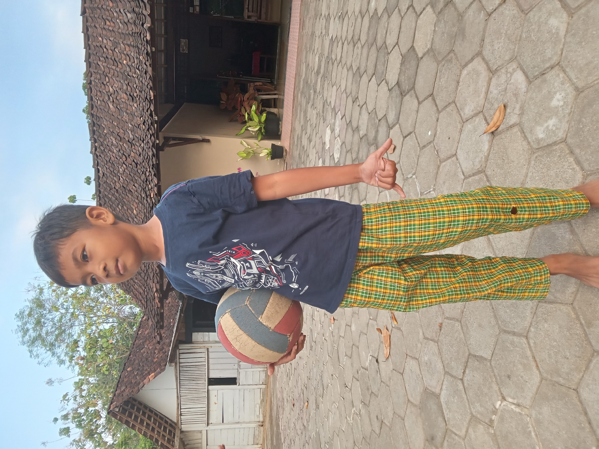 A young boy stands on a hexagonal paver patio, holding a worn, multicolored volleyball in his left arm. He is wearing a navy blue t-shirt with a graphic design and green and yellow plaid pants. His right hand is extended, with his index finger pointing downwards. In the background, a traditional Indonesian house with a tiled roof is visible, with white wooden siding and an open doorway. Potted plants are placed near the entrance. The sky above is clear and blue. The scene appears to be in a rural or semi-rural area.