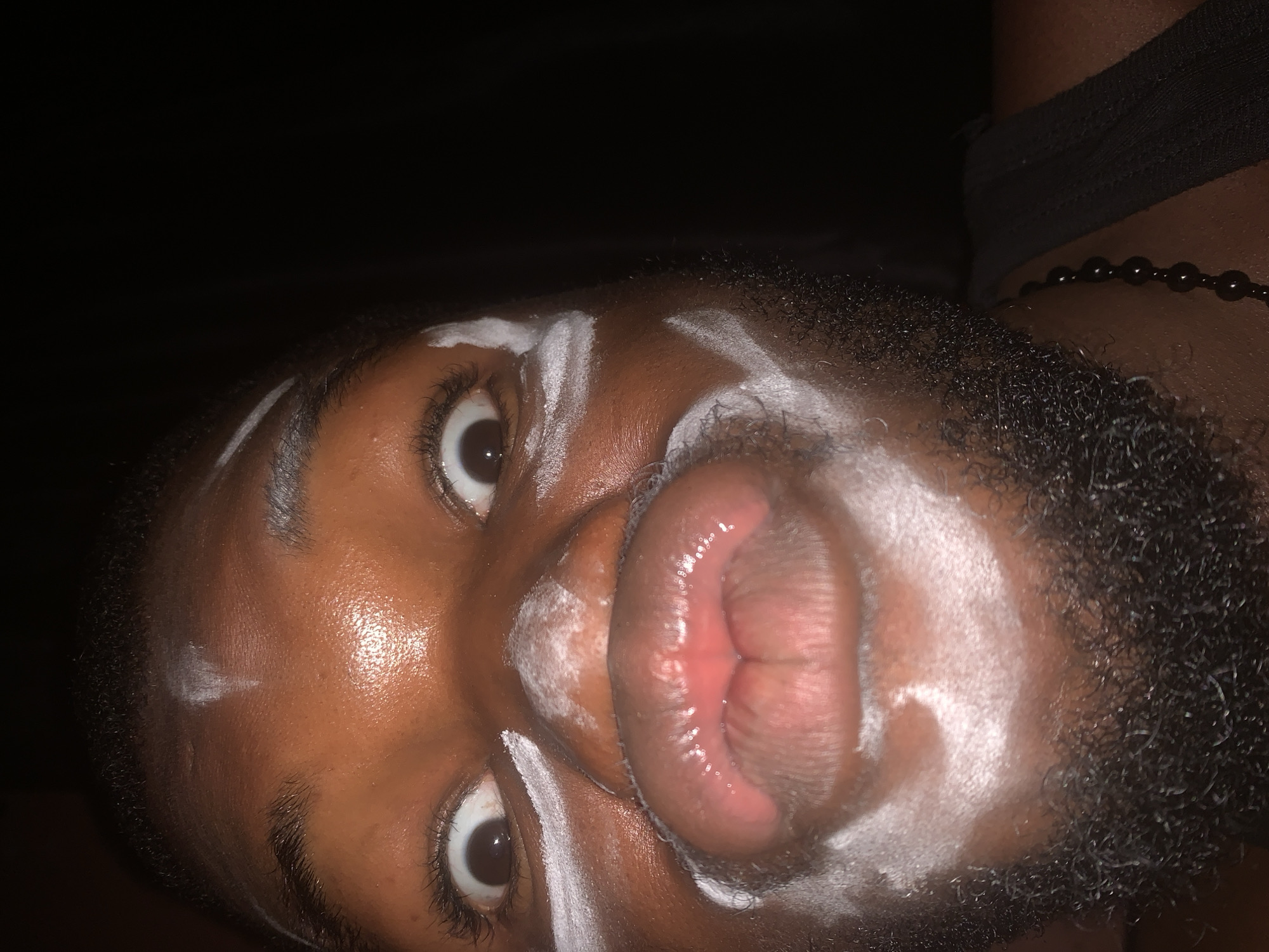 A close-up, rotated portrait captures a person's face against a dark, unlit background, likely taken in low-light conditions at night in Onitsha, Nigeria. The individual has dark skin and prominent white markings painted across their forehead, cheeks, and around their eyes and mouth, forming distinct lines and patterns that suggest body art, ritualistic makeup, or performance adornment. Dark, curly facial hair forms a beard, neatly defining the jawline and chin.
The person looks directly at the viewer with wide, open eyes, revealing dark brown irises and clear whites. Their full, glossy lips are pursed forward into a 'duck-face' or kissy expression, drawing attention to the center of the frame. The skin exhibits a noticeable sheen, reflecting available light. On the right side of the frame, a black beaded necklace is visible against the neck/shoulder, along with a dark fabric strap, possibly part of clothing or a headwrap.
The overall impression is one of playfulness or a stylized presentation, possibly linked to a cultural or celebratory event given the intricate facial markings. There is no visible text in the image.