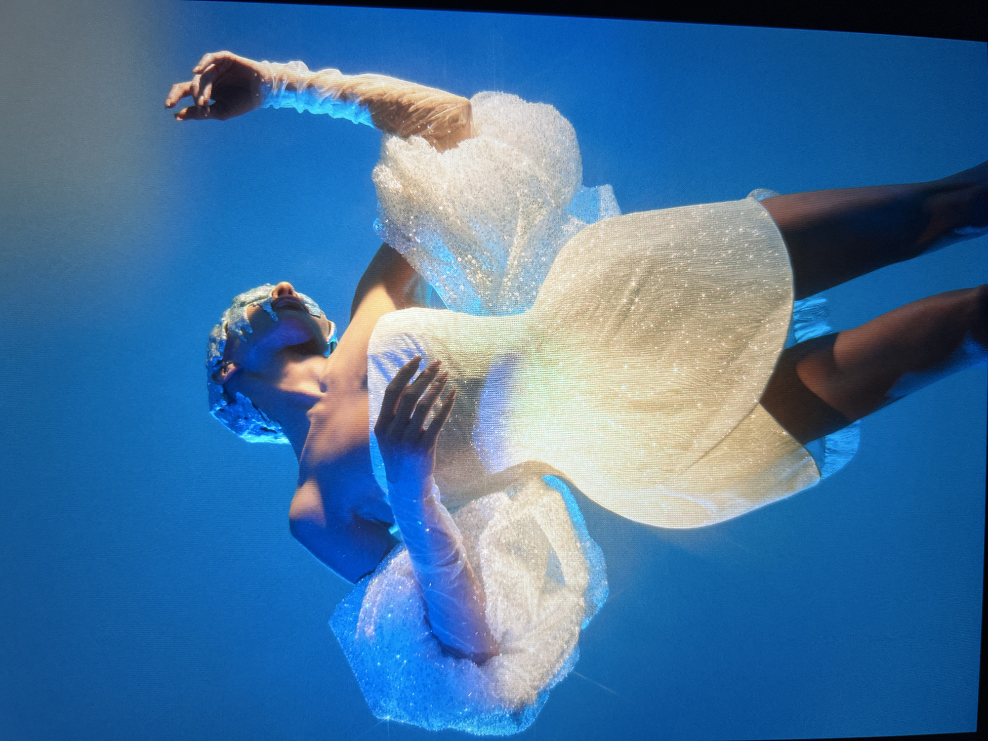 The image shows a person dressed in a sparkling white dress with voluminous sleeves, posing against a blue backdrop. The person's head is tilted back and covered with a shimmering headpiece, and their right arm is raised, with the hand reaching upwards. The dress is short and appears to be made of a sequined fabric. The lighting is dramatic, with bright highlights and shadows. The background is a solid, uniform blue. There is no visible indication of a city or location.