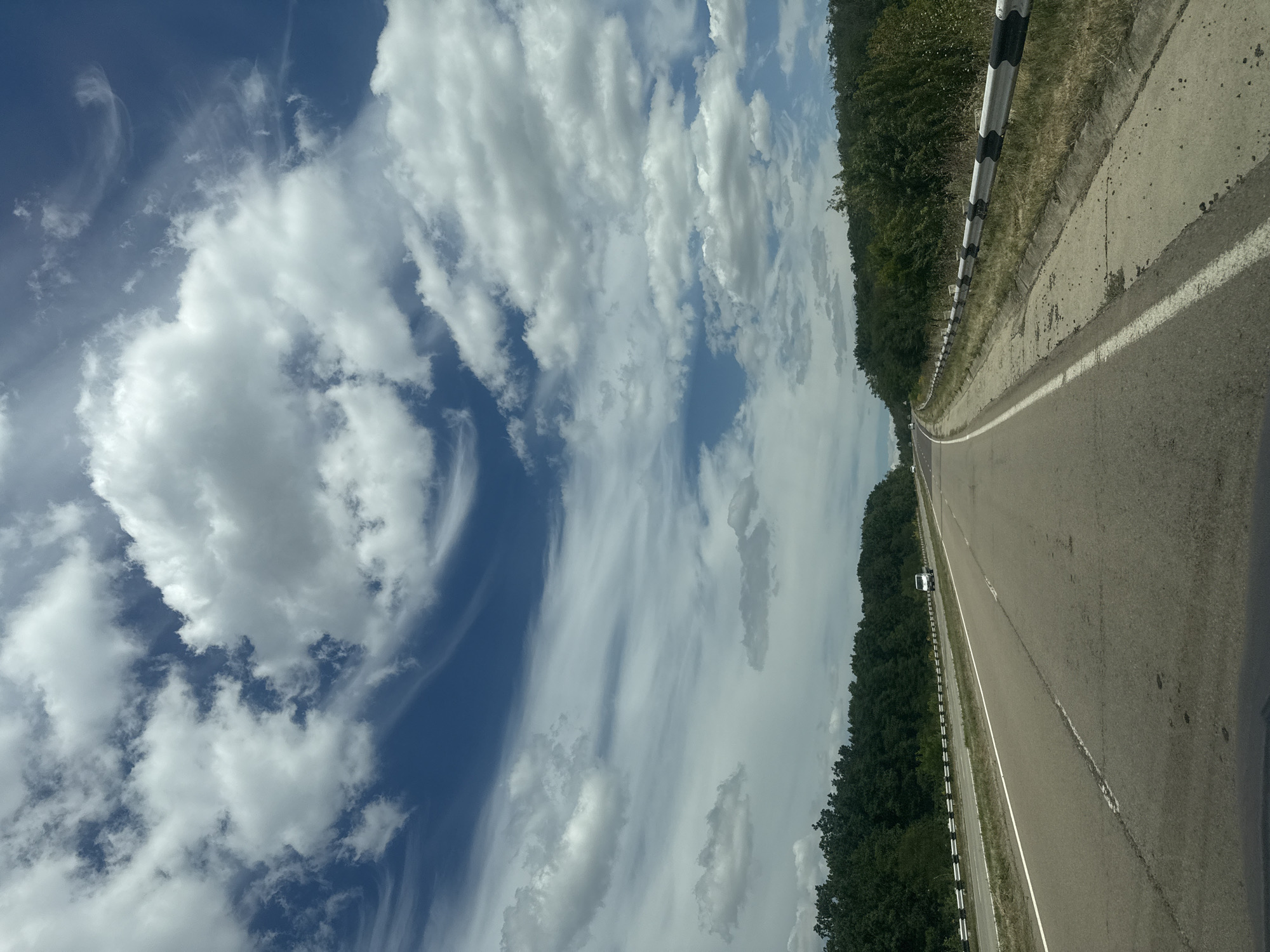 A clear day with a blue sky and scattered clouds is visible over a two-lane asphalt road that curves to the left and disappears into the distance. The road is bordered on both sides by a dense green forest. A white guardrail with black and white markings runs along the right side of the road. A white van is visible in the distance driving on the road. The image appears to be taken from a moving vehicle on the road in Sagaidacul Nou, Moldova.