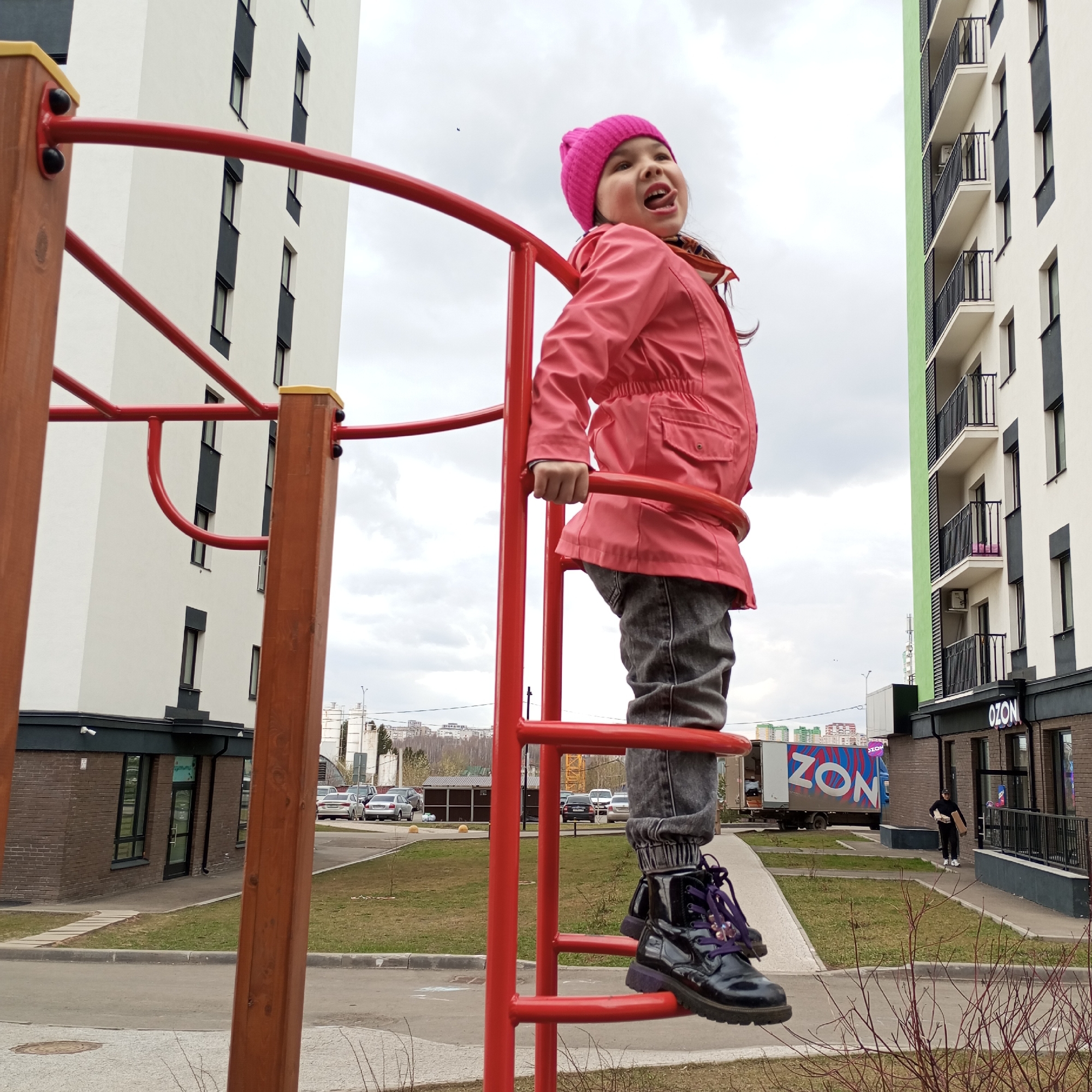 In Pervomaysky, Russia, a young girl with brown hair wearing a pink beanie, a pink jacket, gray pants, and black boots stands on a red playground structure. She has her tongue out and is looking upwards. To the left and right are buildings, and in the background are parked cars and a delivery truck with the text "ZON" on it. A person walks away from the building on the right. The sky is overcast.