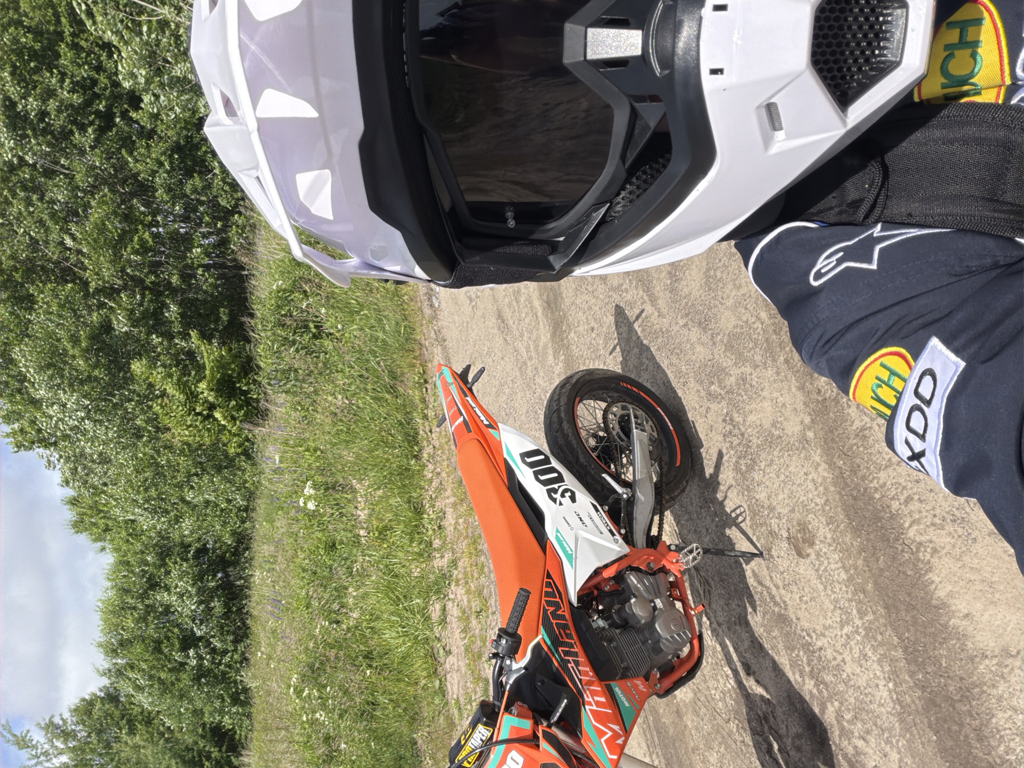 A person wearing a white motocross helmet and a dark blue racing suit is taking a selfie next to an orange and white dirt bike. The dirt bike has the number "300" on its rear. The scene appears to be outdoors on a dirt road, with a background of green trees and a cloudy sky. The location is identified as Povarovka, Russia.