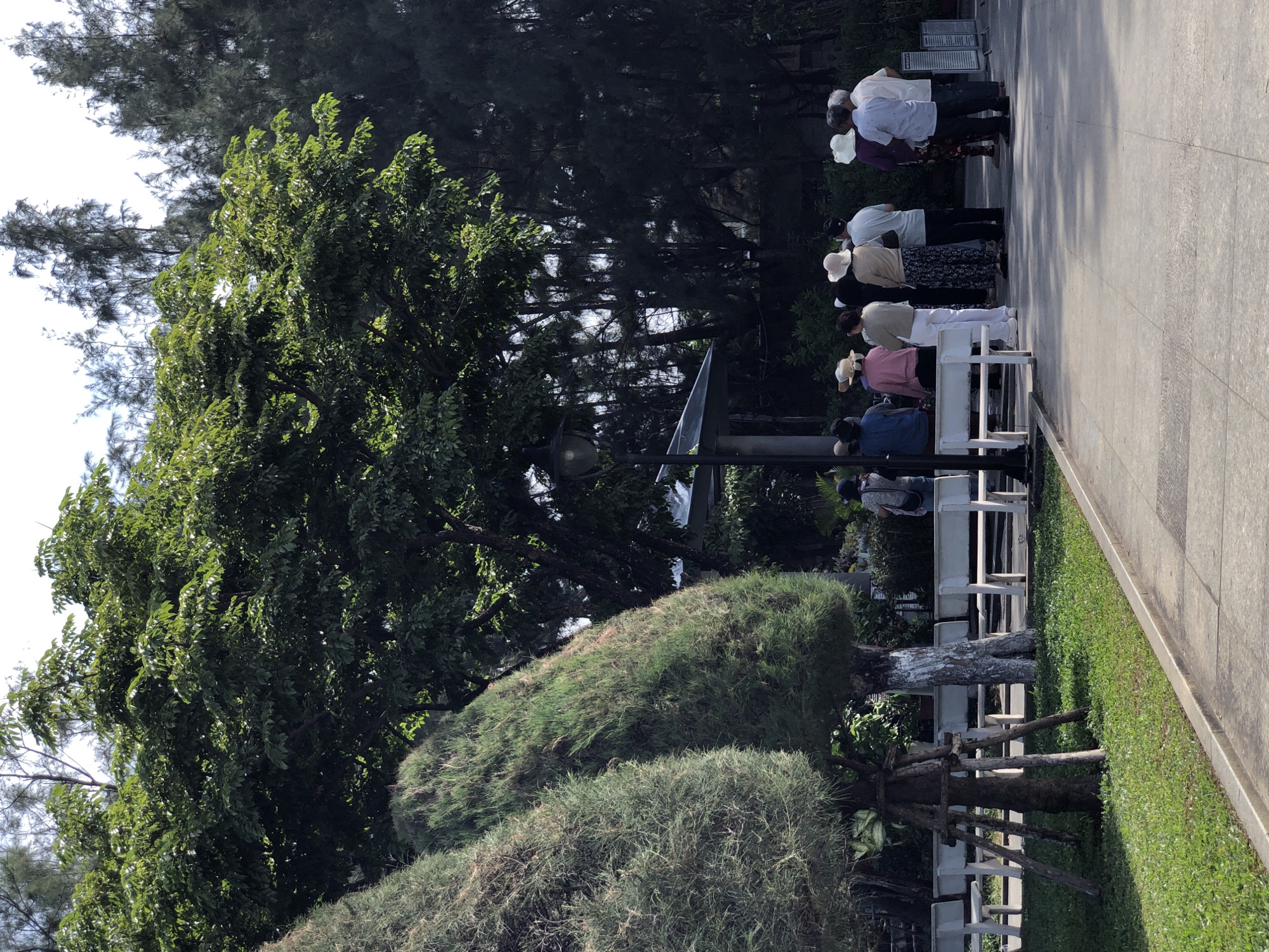 A group of eight individuals, appearing to be tourists, are gathered near a path in a park in Da Nang, Vietnam. The individuals are standing, facing away from the camera, and several are wearing hats. The park features manicured trees, grass, benches, and a lamppost. The overall atmosphere suggests a sunny day.