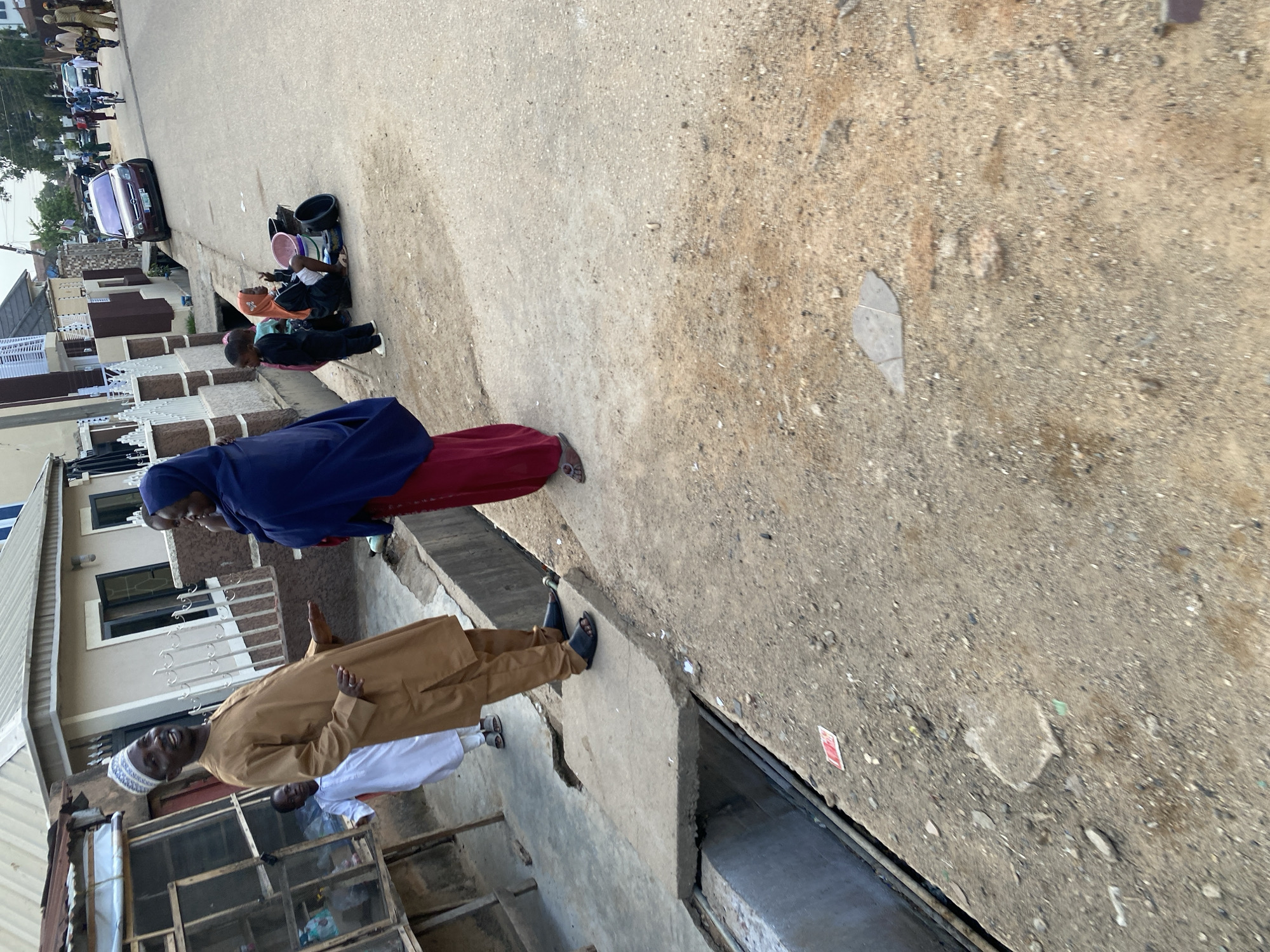 The image depicts a street scene in Jos, Nigeria. In the foreground, a man wearing a brown kaftan and a white cap is smiling and gesturing with his right hand while conversing with a woman. The woman is dressed in a dark blue hijab and a maroon skirt. To their left, a young boy in a white garment stands beside a stall made of wood and metal. In the middle ground, two young children, one in a black outfit and another in an orange headscarf, are sitting on the ground near some buckets and basins. Further in the background, a burgundy sedan is parked on the roadside, and other people and vehicles are visible on the street, suggesting a moderately busy urban environment. The area appears to be a paved street with a concrete drainage channel running alongside it. The overall impression is a slice of everyday life in a Nigerian city.