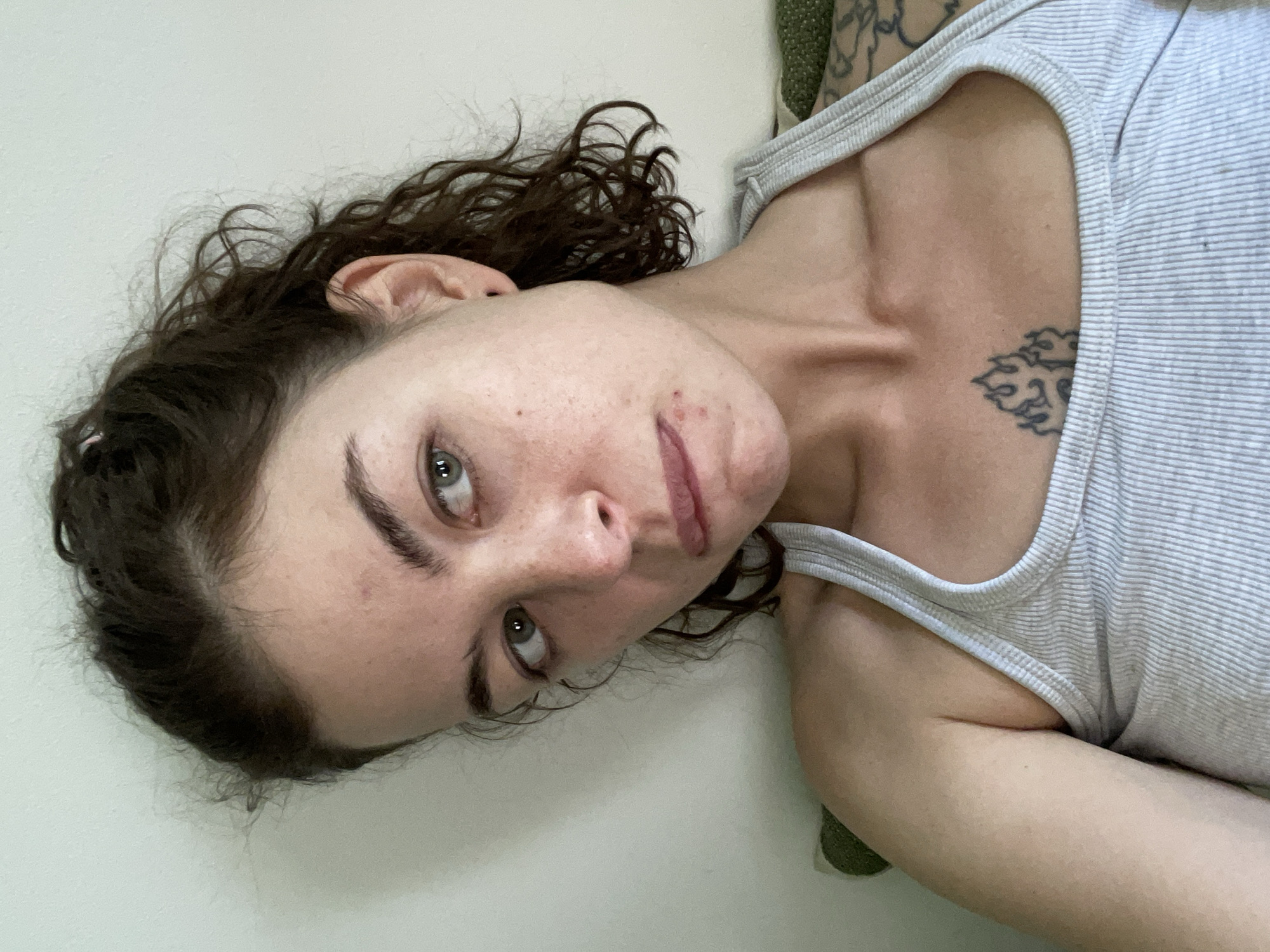 A young woman with curly brown hair pulled back in a messy bun and green eyes is looking off to the side. She is wearing a light gray ribbed tank top. There are some red blemishes on her face, particularly around her mouth. A tattoo of flames is visible on her chest. The background is a plain, light-colored wall. The image is a close-up shot of the woman's upper body.