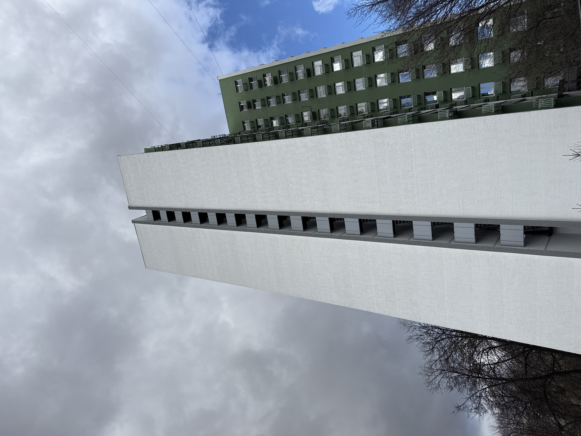 The image depicts the "Izvestia" printing house building, a constructivist architectural landmark located in Moscow, Russia. The view is a low-angle shot, emphasizing the building's height and geometric forms. The building features a large, white rectangular section with a series of recessed, dark openings along its length. Adjacent to this structure is a green building with multiple windows and what appear to be balcony railings. The sky in the background is divided between dark, heavy clouds and patches of blue sky with white clouds. Branches of trees are visible at the bottom and to the right of the frame. A wire stretches across the upper portion of the image.