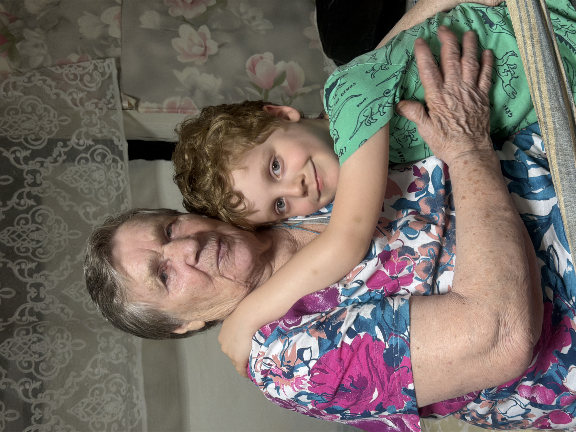 An elderly woman and a young child are closely embracing. The woman has gray hair and is wearing a floral-patterned dress in pink, blue, and white. The child, with light brown curly hair, is wearing a green shirt with dinosaur prints. The woman is holding the child close, with her arm and hand visible. The background includes floral wallpaper in shades of gray and pink, and patterned lace-like fabric. The image's orientation suggests it may have been rotated. The city of Dubrova, Russia is not identifiable from the image.