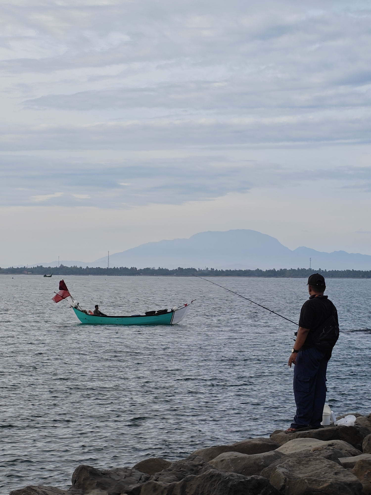 The image shows a coastal scene, likely in Lhokseumawe, Indonesia, given the distinctive geographical features. In the foreground, a man stands on rocky outcroppings along the shoreline, holding a fishing rod extending out over the water. He is wearing dark clothing, including a baseball cap, t-shirt, and pants. He faces toward the ocean. A small white bucket and some plastic are on the rocks near him.
In the mid-ground, a teal-colored fishing boat with a red flag is visible on the water. A person is sitting in the boat. In the background, land is visible. There are mountains that are faded into the distance by haze or fog. The sky is overcast with cloudy formations. The water is choppy with small wave formations.
