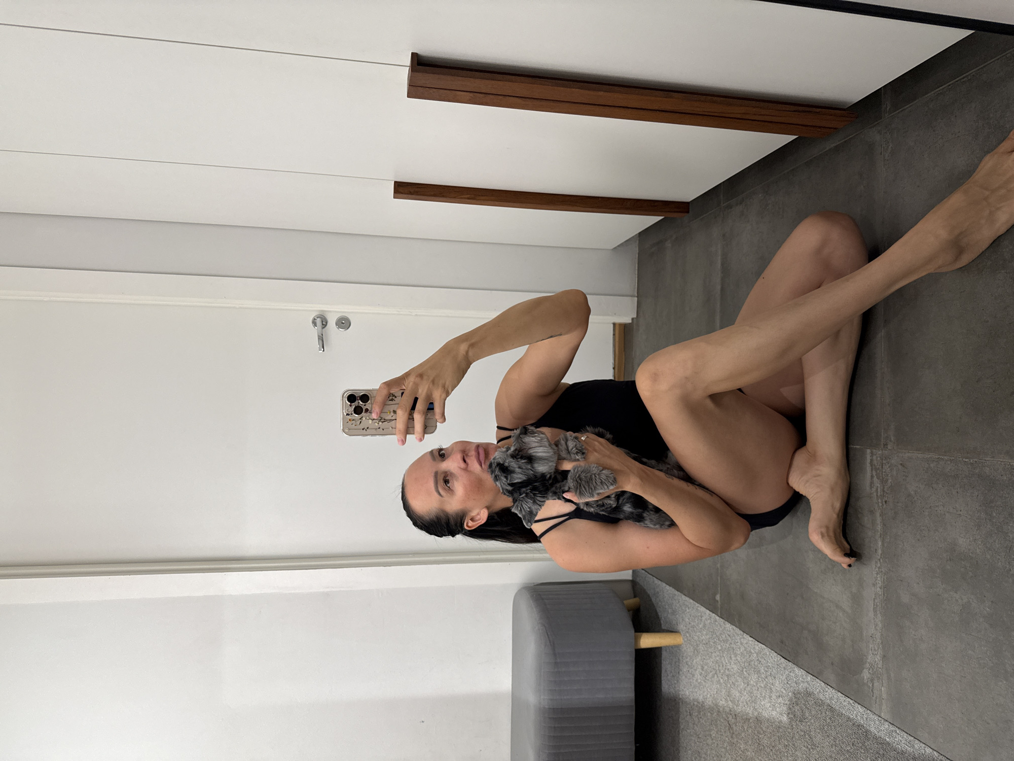 This is a vertical, low-angle shot of a woman holding a small, grey dog in her arms while taking a selfie with her phone. The woman is wearing a black swimsuit and is sitting on a grey tiled floor. Her legs are extended to her right. Her hair is dark and pulled back. She is looking at her phone, which is held in her left hand. The dog, a Miniature Schnauzer, is gray and black with a beard and is nestled in her arms.
In the background, a white door with a silver handle is visible on the left, and a wall with horizontal wooden accents is on the right. A gray, upholstered ottoman with wooden legs is in the lower right of the frame. The lighting appears to be artificial, suggesting an indoor setting. There are no explicit clues about the time of day or weather, but the indoor setting implies it is not an outdoor activity. The overall mood is casual and intimate, with the woman appearing relaxed and focused on her phone and the dog. There is no visible text in the image. The location context of Chaikovsky, Russia is noted, but there are no specific visual cues within the image to confirm this location.