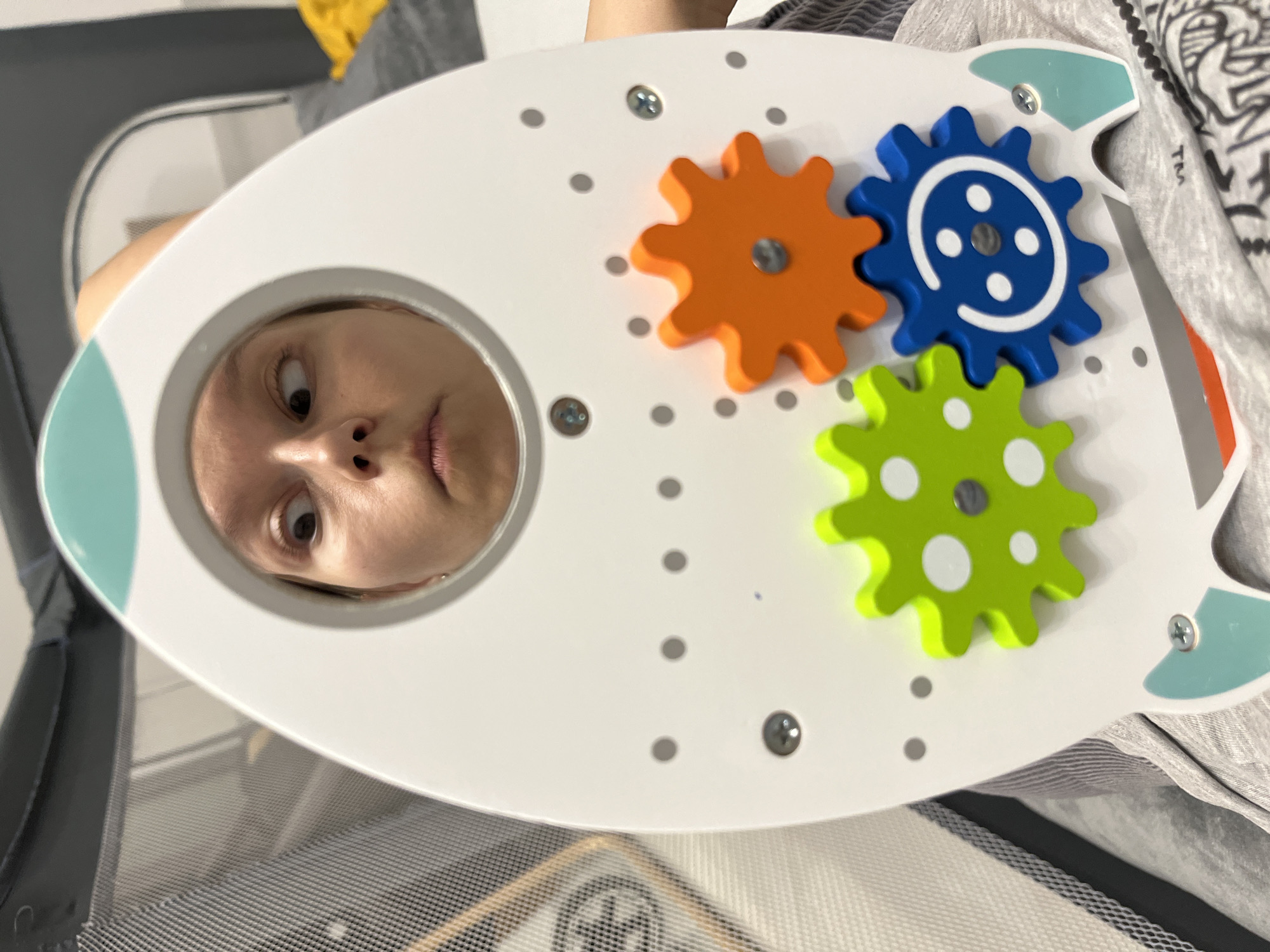 A woman's face, distorted by a circular cutout in a white, rocket-shaped toy with colorful gears, is visible in the foreground. Her eyes are crossed, and her mouth is slightly open. The toy is positioned in front of a person's torso and lap, who is wearing gray clothing with a patterned design. In the background, a mesh crib or playpen is visible. The scene is indoors, with a gray and white patterned matting on the floor and a yellow object in the far background. There is no information available to determine the location of this image within the city of Limassol, Cyprus.
