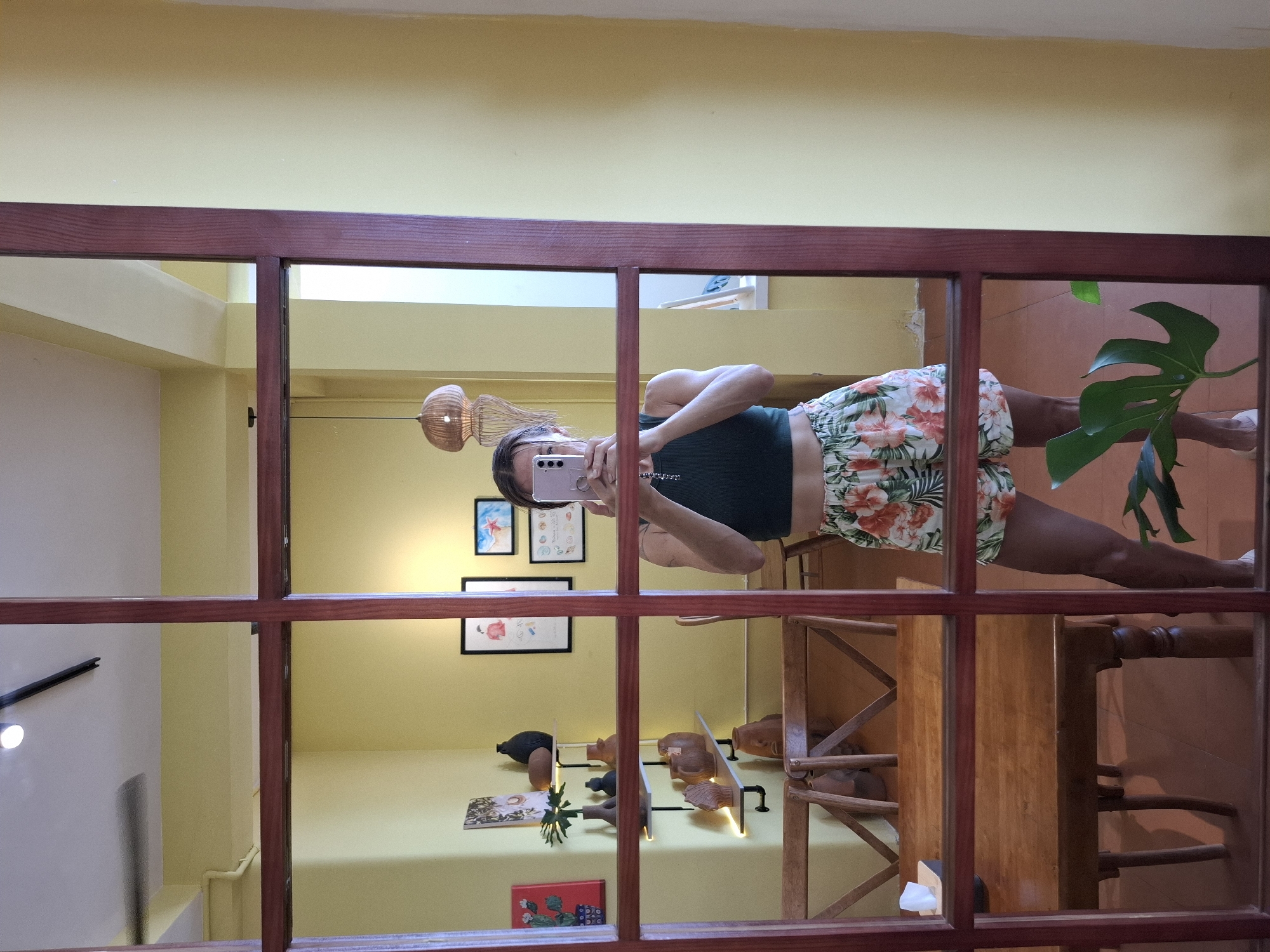 A person is taking a mirror selfie in what appears to be a cafe or shop in Nha Trang, Vietnam. The person is wearing a dark green crop top and floral print shorts. They are holding a pink smartphone to take the picture. The background shows yellow walls, wooden furniture, shelves with decorative items like pottery and artwork, and some hanging light fixtures. The image is framed by a dark wooden partition.