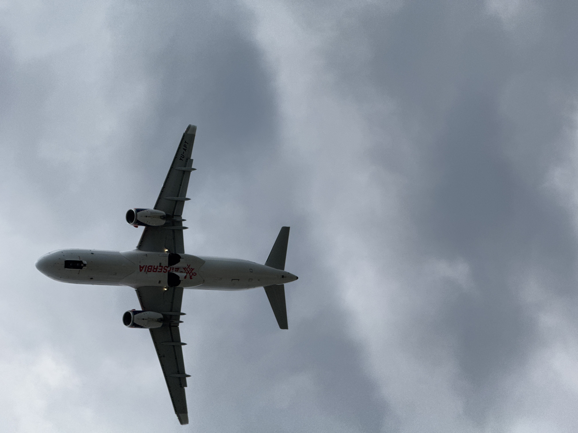 An Air Serbia passenger jet is captured from below as it ascends into a cloudy sky. The aircraft is white with red and blue accents. The word "SERBIA" is visible in red lettering on the fuselage. The registration number "YU-APT" is also present on the wing. The underside of the plane shows its engines, landing gear bay doors, and the tail section. The environment consists of a predominantly gray, overcast sky with visible cloud formations.