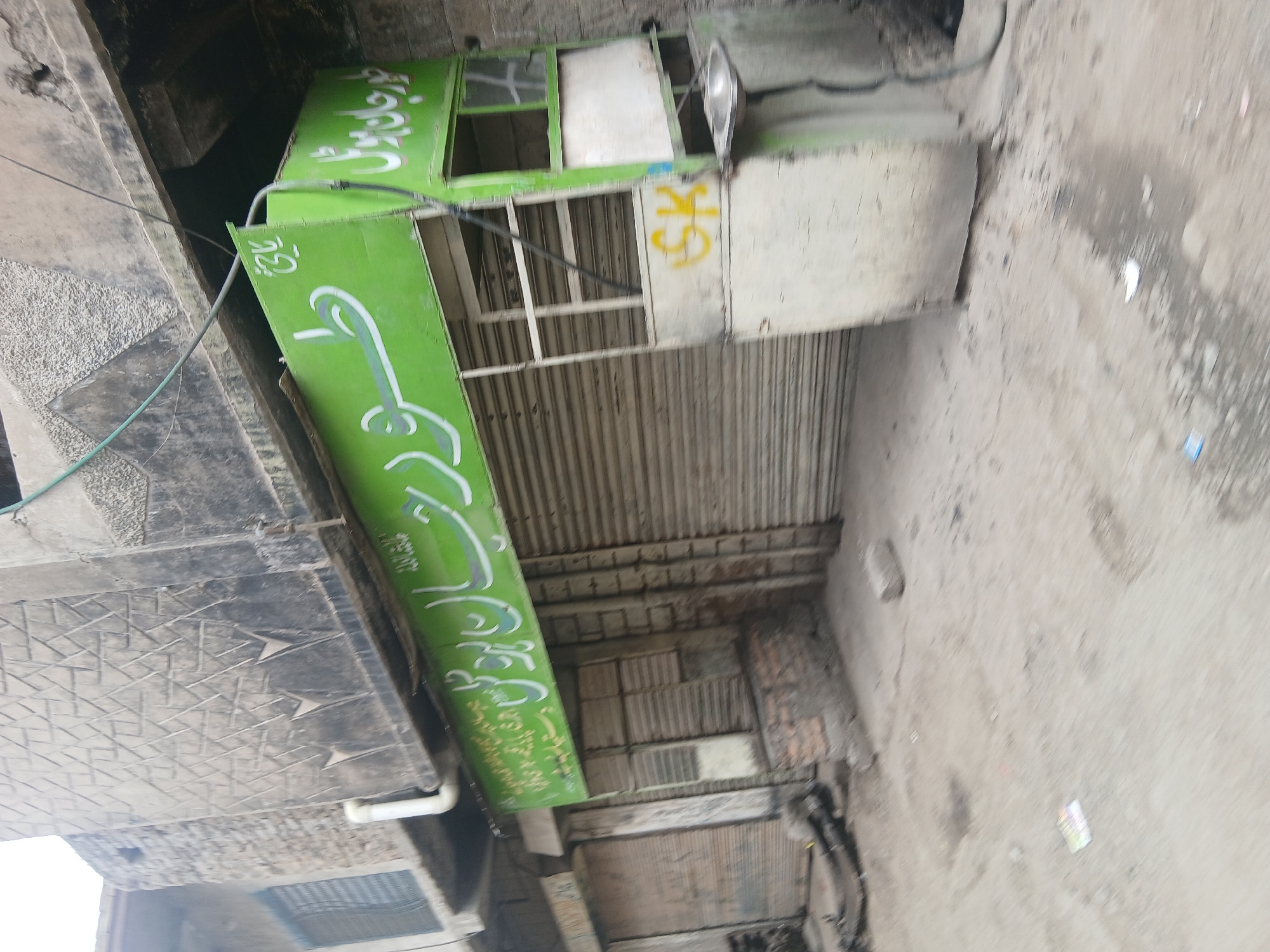 The image shows a closed storefront in Quetta City Tehsil, Pakistan. The storefront is painted green with script on its side. The store has a corrugated metal roll-up door. A weathered light fixture is attached to the side of the storefront. The adjacent street is grey and appears to be wet. The building adjacent to the store is constructed of a dark grey stone or concrete.