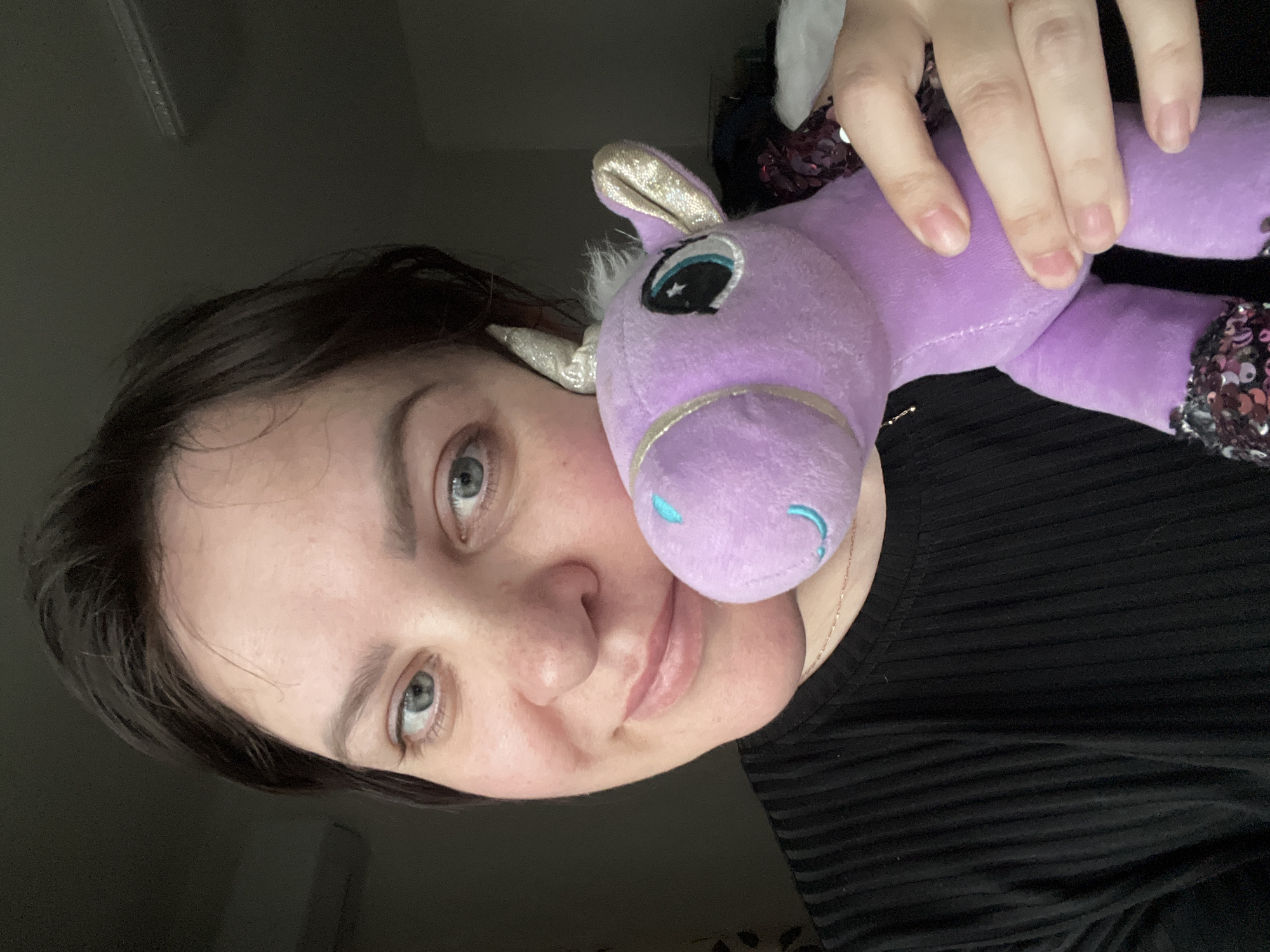 The image, originally captured in portrait orientation but rotated 90 degrees counter-clockwise, displays a close-up of a person holding a purple stuffed unicorn. The subject is a woman with dark hair and striking blue or grey eyes, looking directly at the camera with a subtle, gentle smile. Her skin appears fair, and she is wearing a black, possibly ribbed, top, with a delicate gold chain necklace visible around her neck.
She is holding a soft, purple unicorn or pegasus toy with both hands. The stuffed animal features shiny gold glitter on its ears and horn, a white fluffy mane, and large embroidered eyes with blue irises and small white star accents. A light blue embroidered detail suggests a mouth or nostril on the toy's face. Parts of the unicorn's body, particularly its limbs, are adorned with shimmering silver and purple sequins. The toy is positioned close to the woman's face, almost cheek-to-cheek.
The background is dark and indistinct, suggesting an indoor setting with muted lighting. No specific environmental details or furniture are clearly visible, creating a focused portrait of the woman and her toy. There is no visible text in the image. The activity appears to be posing for a casual photograph, possibly a selfie. The overall mood is calm and friendly. The location context provided is Perm, Russia.