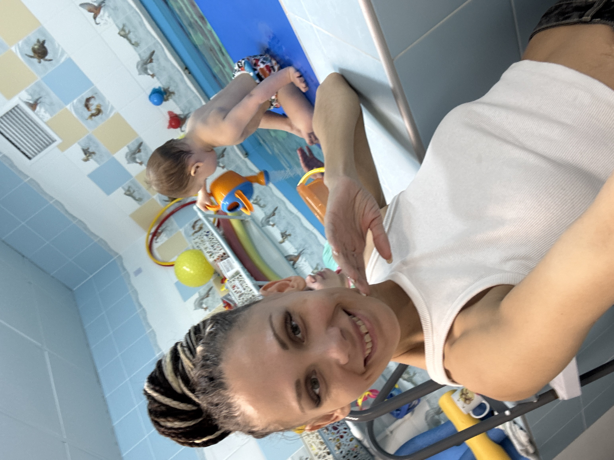 In a bright, presumably indoor setting, a woman with braided hair smiles towards the camera, with a child playing in a blue tub behind her. The child, wearing swimming trunks, is engrossed with a toy watering can. The scene is adorned with colorful tiles and water-themed decorations on the walls. The child and the woman are likely in the city of Rusanovo, Russia.