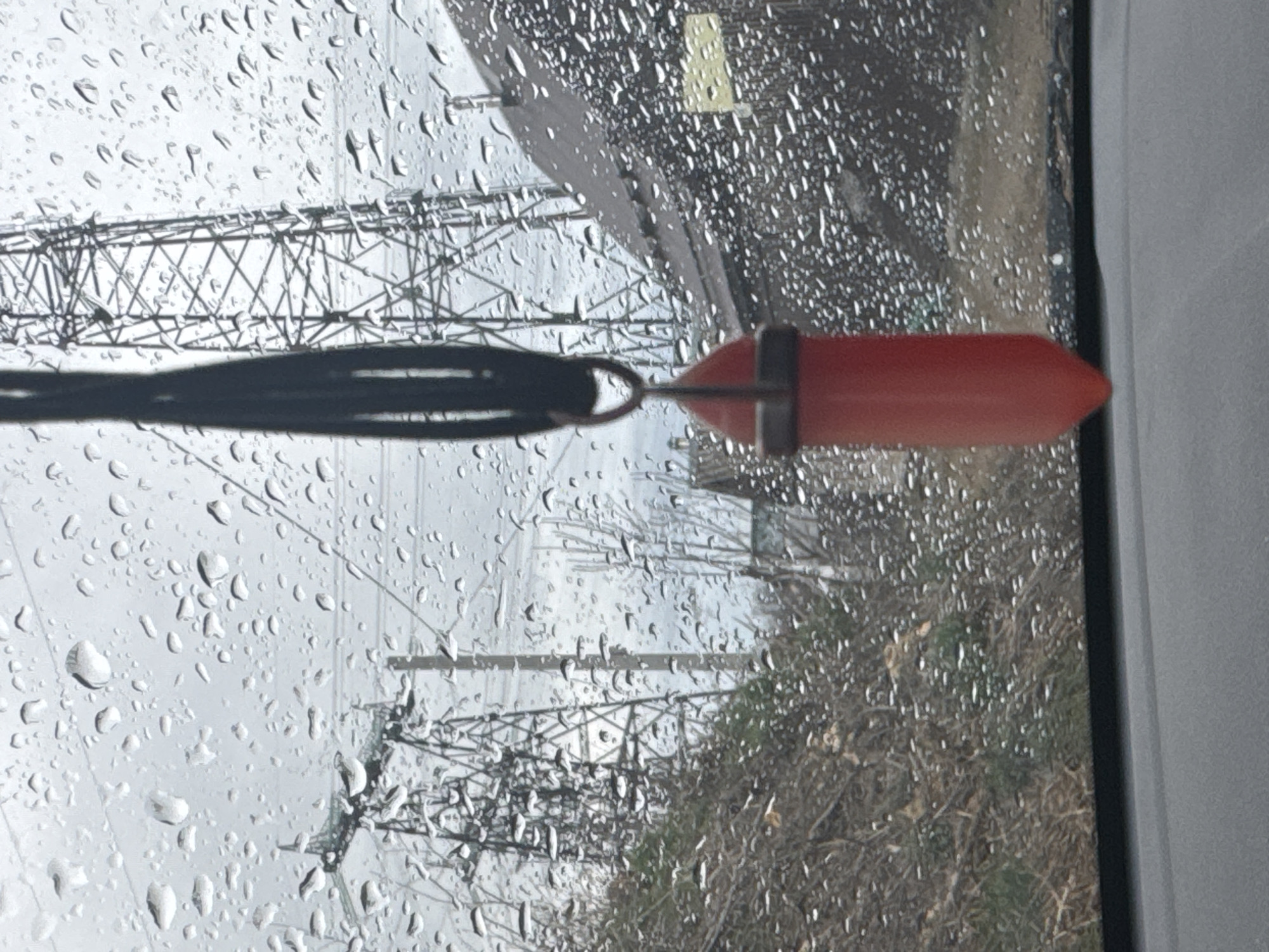The image, taken through a rain-covered window, shows a red, torpedo-shaped object dangling from a black cord. Outside the window, the scene includes high-voltage power lines and a partially visible landscape of vegetation. Given the reference to the city of Chaikovsky, Russia, the setting is likely an urban or semi-urban environment where such infrastructure is present. Specifics about people, actions, or detailed interactions are not discernible from the provided image. The dominant characteristics are the wet window, the hanging object, and the industrial-adjacent scenery beyond.
