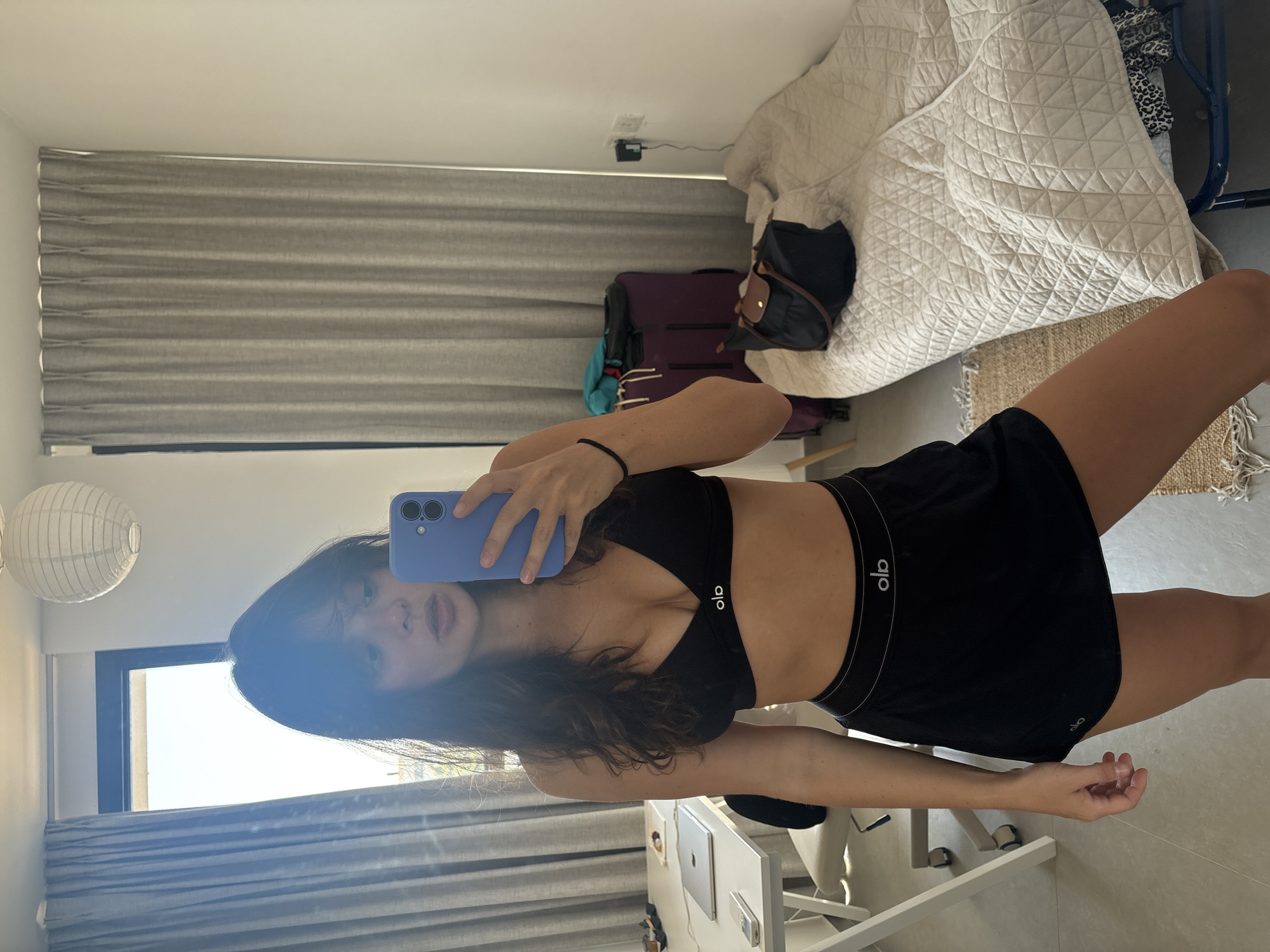 A young woman with dark, wavy hair stands in a room, taking a selfie with a purple smartphone in her right hand. She is wearing a black sports bra with a white logo and black athletic shorts with a white logo. Her left hand is on her hip. The room appears to be a bedroom or living space, with a bed covered in a quilted off-white comforter on the right side of the image. A dark blue suitcase and a black duffel bag are placed on the floor near the bed. On the left side of the image, a white desk with a laptop and a phone is visible. Behind the desk, gray curtains are drawn over a window. A white paper lantern hangs from the ceiling. The flooring is tiled. The overall scene suggests a casual, possibly temporary living space in Tel Aviv, Israel.