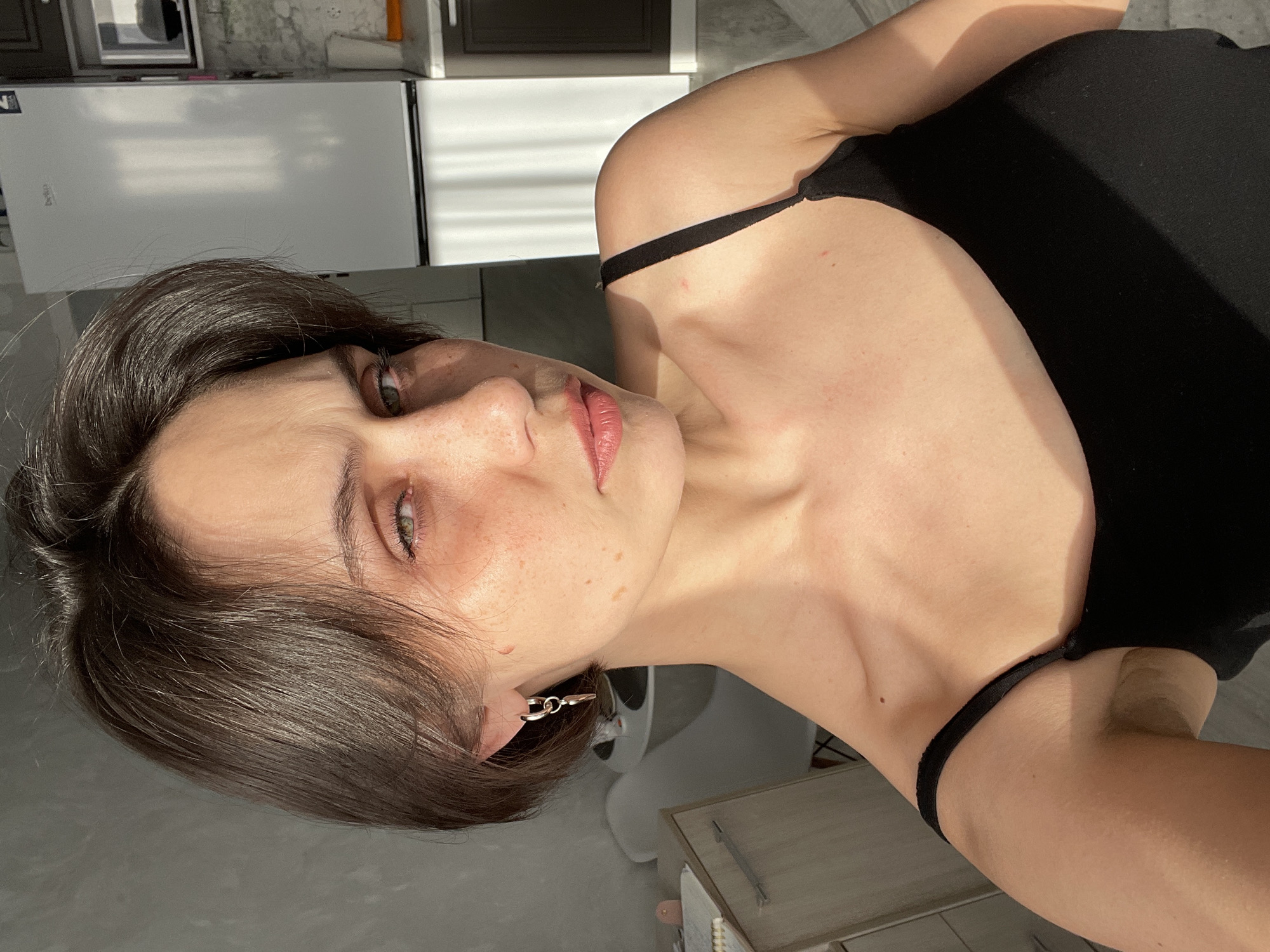 The image is a sideways, close-up portrait of a young woman with short, dark brown hair and light green or hazel eyes. She has fair skin with visible freckles across her nose and cheeks. Her lips are a light pink color. She wears a simple black top with thin straps, and a small, silver-toned hoop earring is visible on her left ear.
The woman is looking directly at the camera with a calm, neutral expression. Strong, natural sunlight illuminates her from the side, creating distinct shadows on her face, neck, and shoulders, and highlighting her hair. The lighting suggests it is daytime, possibly morning or late afternoon.
The background indicates an indoor setting, likely a domestic kitchen or a room adjacent to one. A white refrigerator, which appears to be a "Beko" brand (text faintly visible), is seen behind her, along with kitchen counters or cupboards. The surfaces in the background show reflections, possibly from a window or another light source. The overall impression is a casual, sunlit moment captured in a home in Izhevsk, Russia.