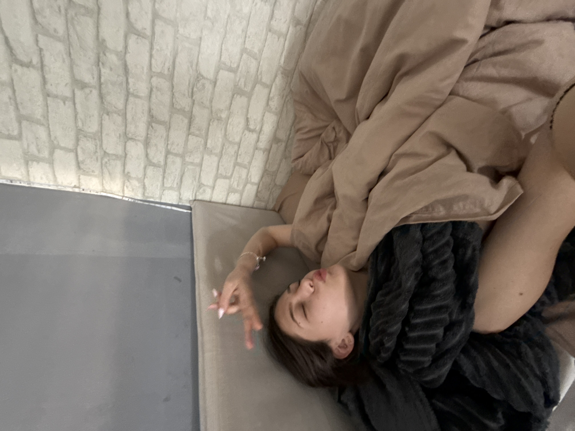 The photograph shows a woman lying on a bed or sofa in a room with a brick-patterned wall. She is partially covered by a beige duvet and a dark gray, textured blanket. Her face is visible, and she is wearing a bracelet on her wrist. No specific details indicate the location is in Sofia, Bulgaria. The scene suggests a relaxed, indoor setting.