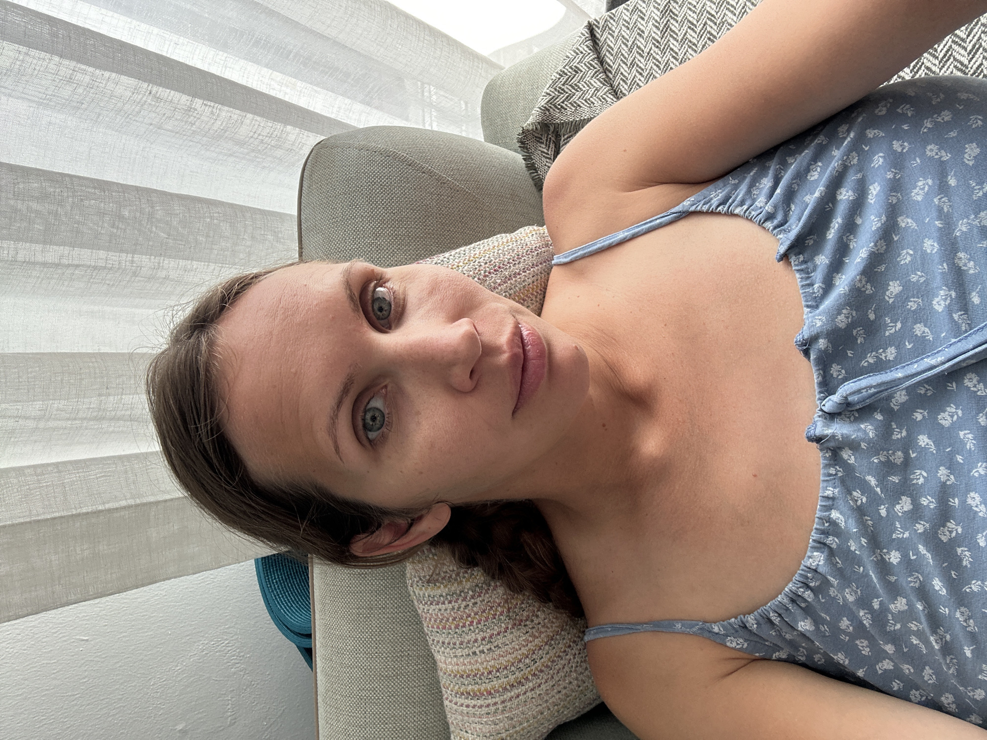 A fair-skinned woman with light brown hair and light eyes is reclining on a sofa. She is wearing a light blue floral-patterned top with thin straps. She is lying on a patterned pillow. A grey sofa is behind her, and a set of white horizontal blinds is in the background. The location cannot be determined based on the image.