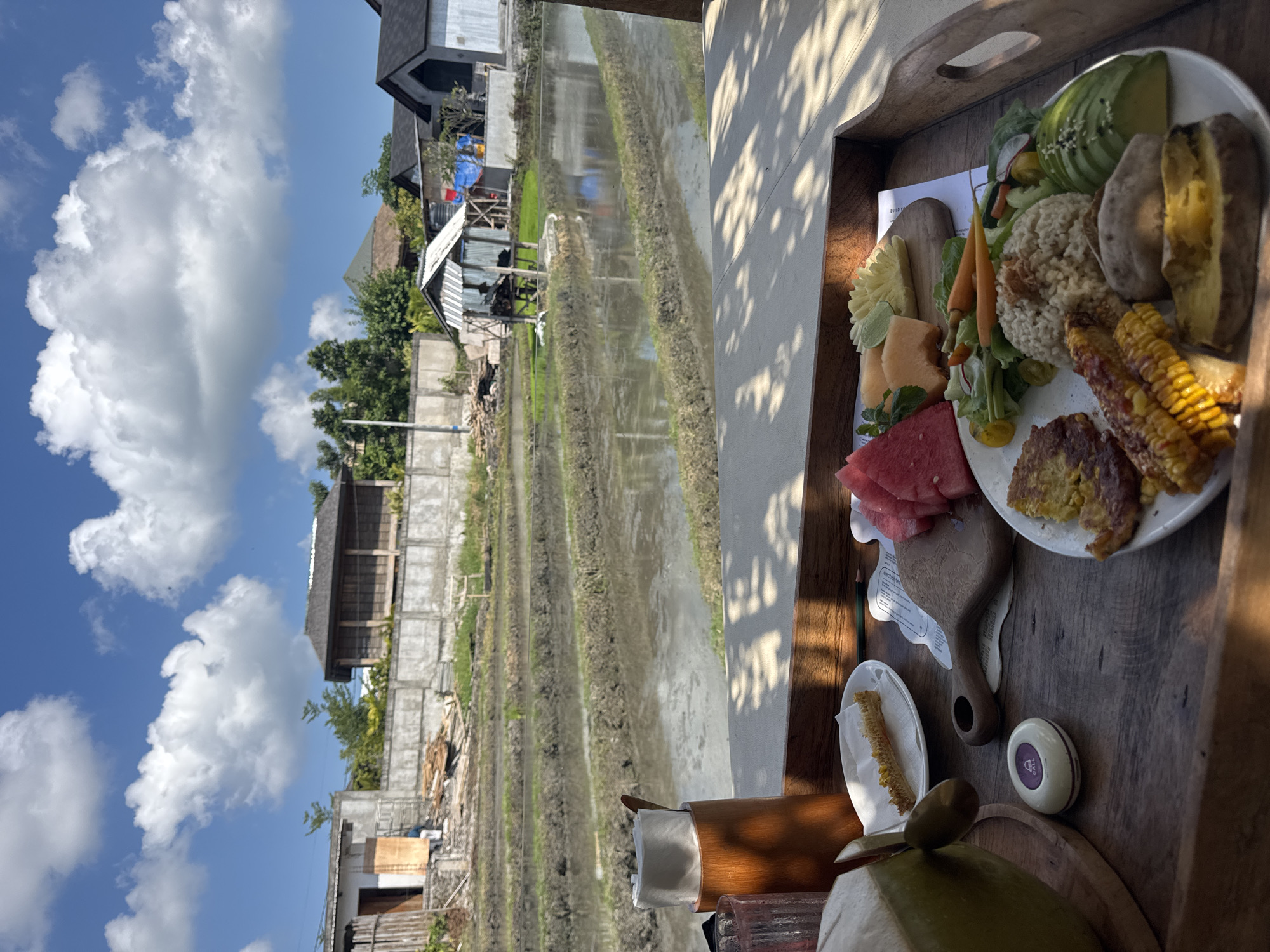 An outdoor scene in North Kuta, Indonesia, captures a diverse meal served on a wooden tray, overlooking a landscape of rice paddies under a bright blue sky.
In the foreground, a wooden tray holds a substantial meal. A white plate features sliced avocado, a mound of cooked grain (possibly brown rice), two cooked sweet potatoes, two pieces of grilled corn on the cob, several savory fritters or patties, and a small salad with greens and baby carrots. Adjacent to the plate, a small wooden cutting board displays an assortment of fresh fruit, including slices of red watermelon, melon wedges, and carved pineapple. Other items on the tray include a green young coconut with a straw for drinking, a wooden holder containing white napkins, a small dish with a segment of corn cob, a pencil, and a white and purple circular button clearly labeled "CALL." Partially visible text on a white paper beneath the fruit board reads "electr" and "services."
Beyond the dining setup, the mid-ground is dominated by neatly structured rice paddies, which appear to be either flooded or recently tilled, with water reflecting the sky. These paddies stretch horizontally across the frame. In the background, beyond the paddies, several houses and buildings with various architectural styles are visible, interspersed with green trees and some apparent construction or debris.
The scene is set on a clear, sunny day, indicated by the strong natural light and distinct shadows cast on the wooden tray and an adjacent wall. Large, fluffy white cumulus clouds are scattered across the blue sky. The overall atmosphere suggests a peaceful, rural setting, highlighting a meal enjoyed amidst the natural beauty of the agricultural landscape.