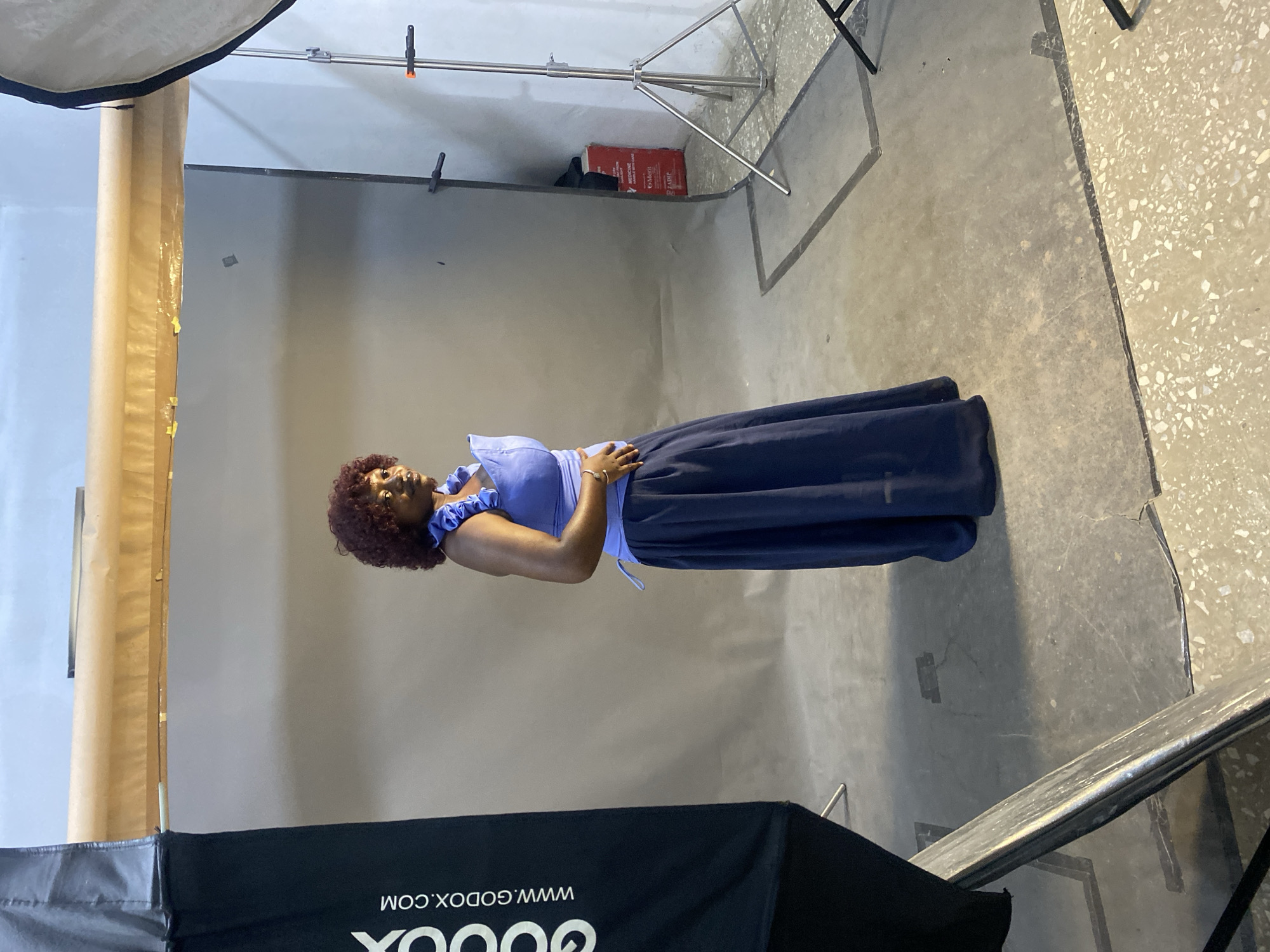 In Jos, Nigeria, a woman with curly red hair and brown skin poses in a studio. She is wearing a blue ruffled top and a long, dark blue skirt. The backdrop is a plain gray wall, with a paper roll suspended above it. To the left, a black softbox with "GODOX.COM" printed vertically is visible. To the right, a metal stand with a white softbox is positioned, and a red box with white lettering that appears to say "MEDICINE" is also present. The floor is concrete.
