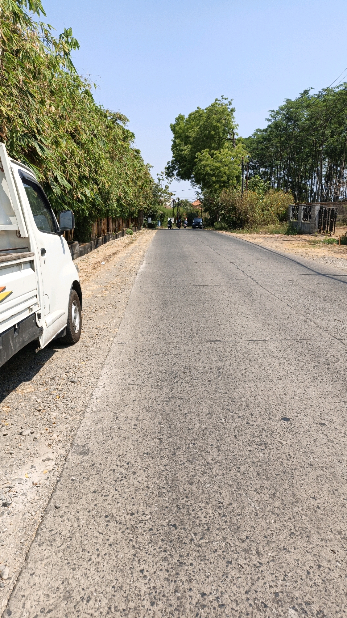 In Bandengan, Indonesia, a white utility vehicle is parked on the left side of a paved road. The vehicle is positioned at an angle, facing away from the camera and towards the distance. A line of lush bamboo trees borders the left side of the road, separated by a low fence. The road stretches into the distance, where several motorcycles and a car are visible. The right side of the road is lined with trees and vegetation, and a small concrete structure with a metal grille can be seen. The sky above is clear and blue.