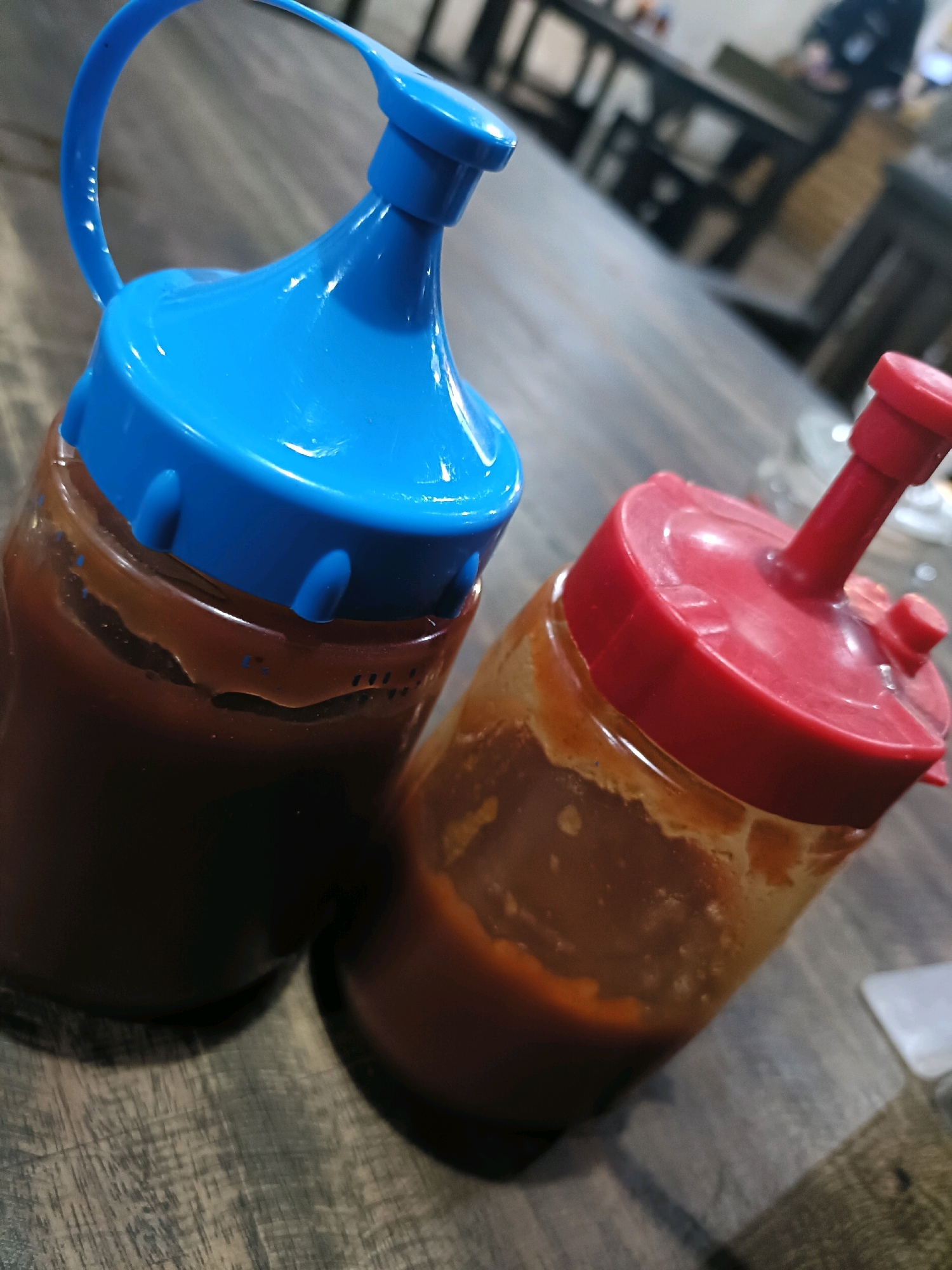 Two sauce bottles with blue and red caps sit on a wooden table. The bottle on the left has a blue cap and contains a dark brown sauce. The bottle on the right has a red cap and contains a lighter brown sauce. The bottles are positioned close together, with the blue-capped bottle slightly in front of the red-capped one. The wooden table has a textured surface with visible grain. In the background, out of focus, are tables and chairs, suggesting a restaurant or food establishment setting. A person is dimly visible in the far background, appearing to be seated at a table. The overall lighting suggests an indoor environment.
