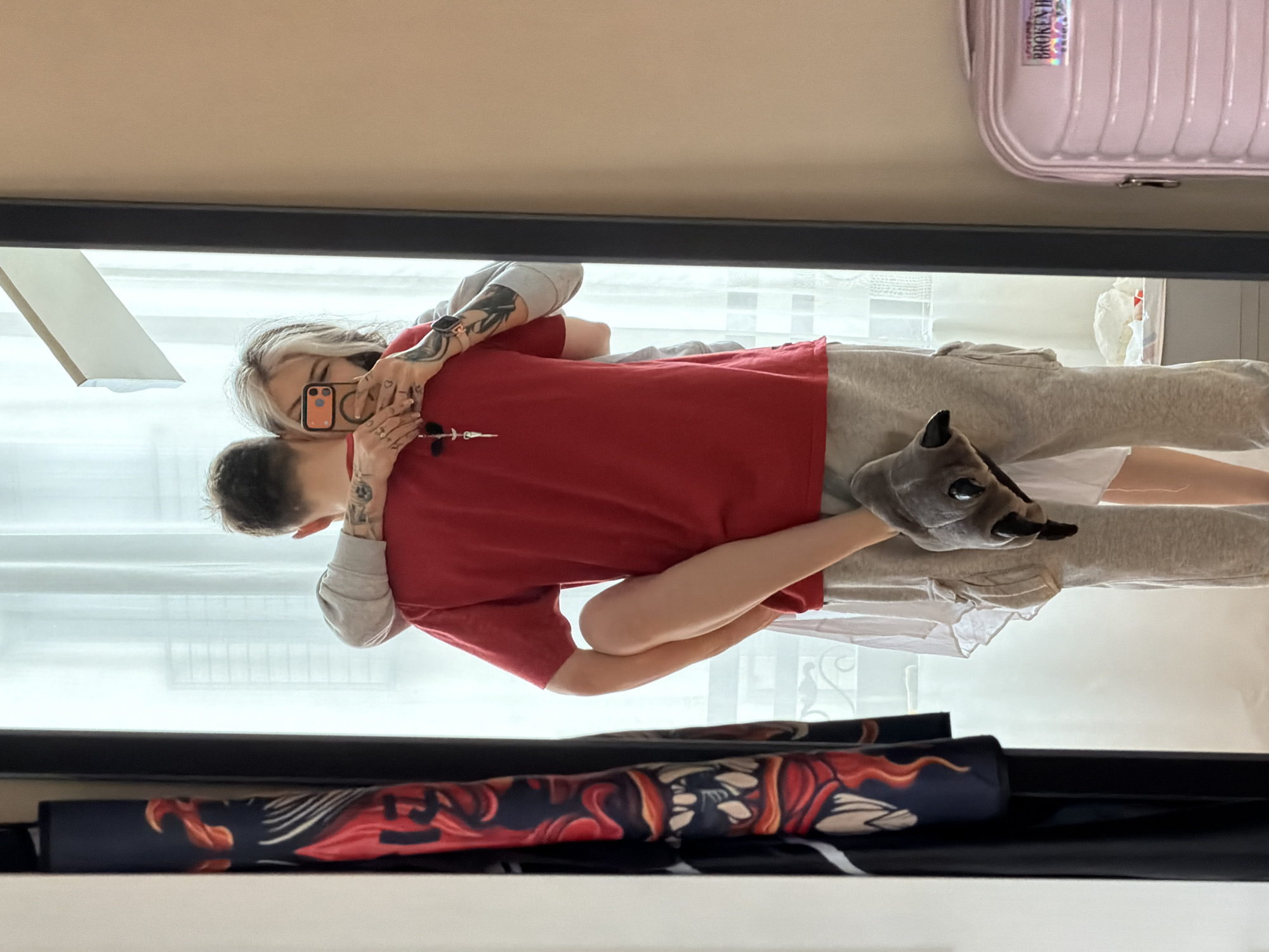 The scene depicts two individuals embracing in an indoor room, potentially a residence or hotel room in Budva, Montenegro, though specific city landmarks are not visible. The image is a mirror selfie.
The individual in the foreground, facing away from the camera, appears male. He wears a red short-sleeved t-shirt and light grey sweatpants. Tattoos are visible on his upper arms/shoulders. A silver-colored necklace with a cross-like pendant hangs around his neck.
The individual embracing him, reflected in the mirror and facing the camera, appears female. She has two-toned hair, dark at the roots and blonde/white at the ends. She wears a light grey long-sleeved top and a white garment (possibly a dress or skirt) underneath, partially visible. Tattoos are present on her forearms, and a watch is on her left wrist. She holds a smartphone with an orange case and a visible circular attachment (MagSafe ring) in her right hand, taking the mirror selfie. Her left arm is wrapped around the first individual's neck, and her right arm around his waist. Her left leg is lifted, with her bare foot, clad in a grey plush animal-paw-shaped slipper, resting on the first individual's left hip/thigh area.
The background includes a large rectangular mirror with a black frame. To the left of the mirror, a black fabric with a detailed red and orange fiery or dragon-like pattern is hanging. Behind the embracing individuals, a window covered by sheer white curtains is visible, allowing diffused light into the room. To the far right, partially reflected in the mirror, is a light pink hardshell suitcase with a "BROKEN HEARTS" sticker on it. The room's walls are a plain, light color.