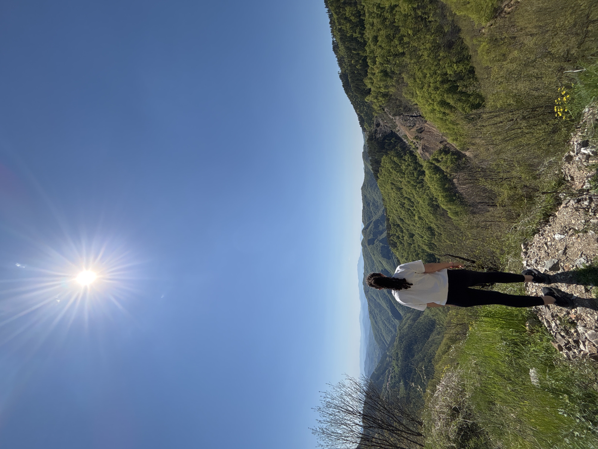 The image shows a woman standing on a mountain edge in Petrov Dol, Bulgaria. She is wearing a white shirt and black leggings. The mountain range is visible in the background, with trees and vegetation. The sky is blue and the sun is shining brightly.