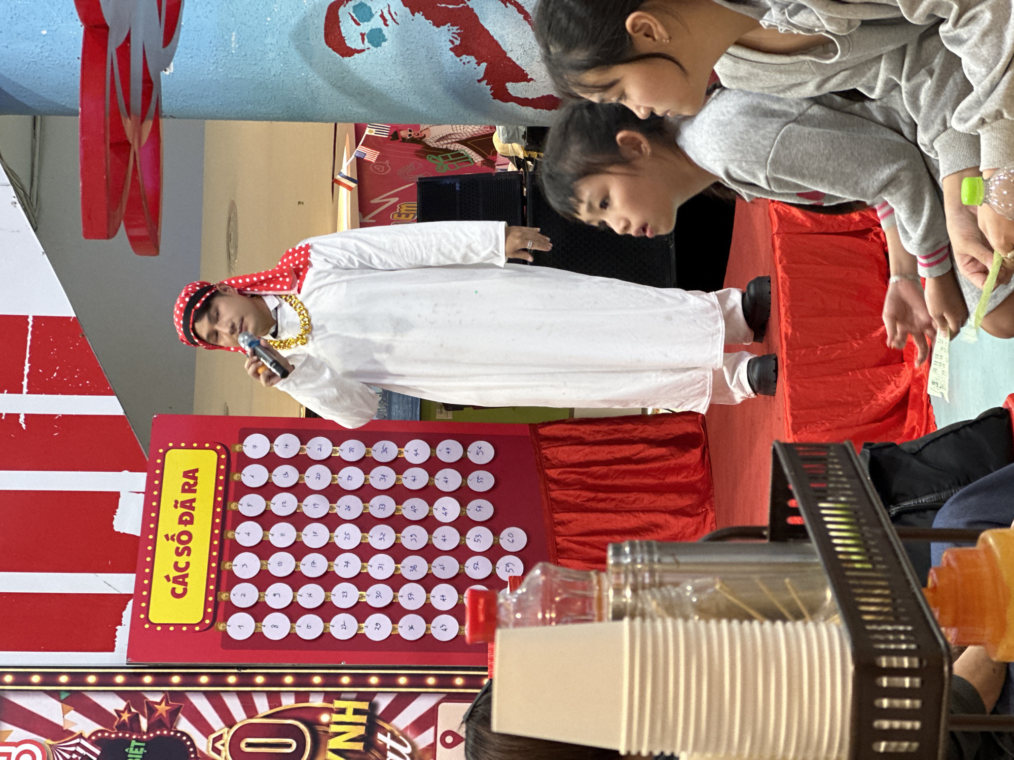 This image captures a lively event, likely a lottery or game show, taking place outdoors in Da Lat, Vietnam. The scene is dominated by vibrant red decorations and a stage setup. In the center, a man dressed in traditional Middle Eastern attire, including a white thobe and a red and white patterned headscarf, holds a microphone, appearing to be the host or presenter. He wears a prominent gold chain.
In front of him is a large red board with numerous circular slots, each containing a number. The text "CÁC SỐ ĐÃ RA" is prominently displayed in a yellow box on this board, translating to "Numbers Drawn." A grid of numbered circles, from 1 to 55, is visible, some of which appear to have been selected or marked.
To the right of the presenter, two young girls are leaning over the stage, observing the proceedings with focused expressions. They are dressed casually in grey hoodies. The setting suggests a public gathering or a festive occasion, possibly during the day given the bright lighting. The overall atmosphere appears energetic and engaging, with elements of suspense and excitement implied by the game board and the attentive audience. In the foreground, out-of-focus plastic cups and a black metal dispenser suggest concessions or a drink station. The presence of Vietnamese text on a banner at the bottom, partially obscured but reading "BIỆT" and "NH", further confirms the location.