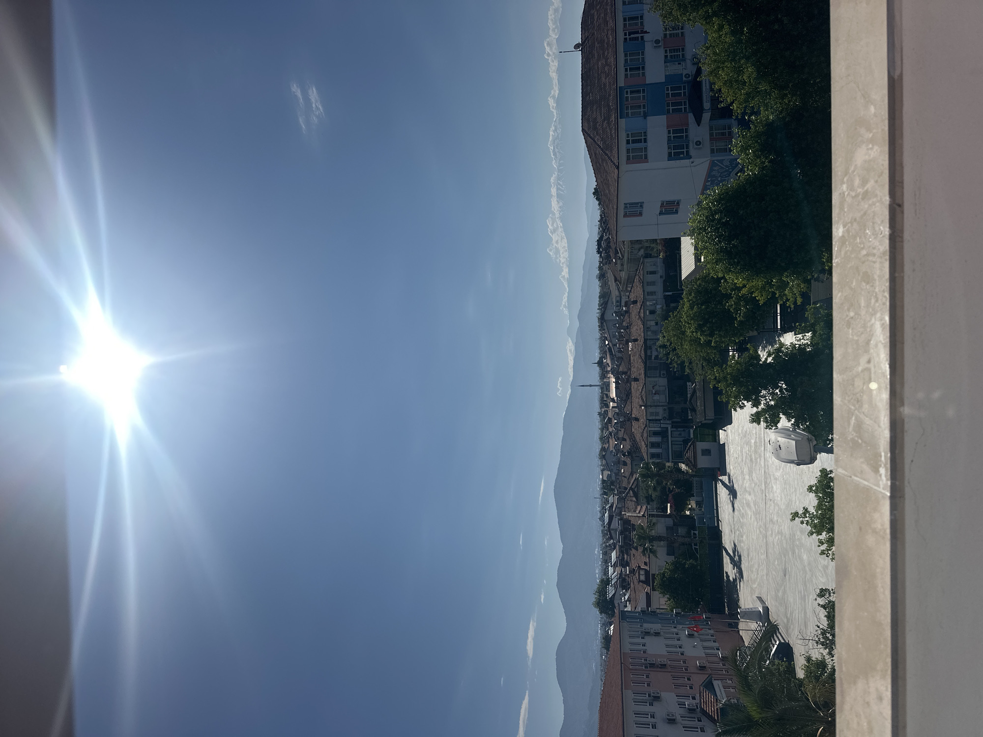 The image shows a view of Fethiye, Turkey, from an elevated position. The sky is bright blue with a few scattered clouds and a strong sun glare to the left. The urban landscape includes buildings of varying sizes and styles, with a mix of residential and commercial structures. Trees are interspersed throughout the scene, adding greenery to the urban environment. A large, open paved area is visible, likely a public square or plaza. In the distance, mountains can be seen. A prominent minaret is visible. The buildings appear to have air conditioning units. A window frame is visible on the right.