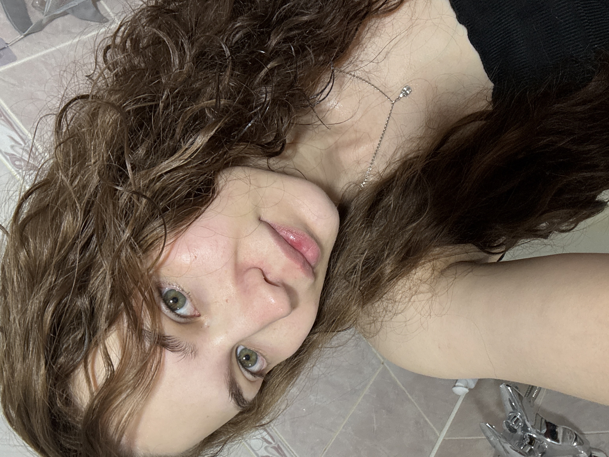 An overhead close-up shot features a young person, likely female, lying down and looking directly at the camera. Her head is angled towards the bottom left of the frame, with her body extending diagonally towards the top right. She has long, wavy, dark brown hair that appears damp or wet, with individual strands visibly defined. Her complexion is fair, and her eyes are a striking green. Her lips are slightly parted, revealing a natural pink hue, and her expression is calm and direct.
She is wearing a dark-colored (possibly black) top or strap, visible on her upper body and shoulder. A delicate silver necklace with a small, sparkling pendant or bead hangs around her neck, draped across her collarbone. The background consists of light-colored, possibly beige or grey, tiled surfaces, suggesting an indoor setting like a bathroom. In the bottom right corner, a chrome-plated fixture resembling a shower head or faucet handle is partially visible, further supporting the bathroom environment. The dampness of her hair suggests she may have recently showered or bathed. No visible text is present in the image.
