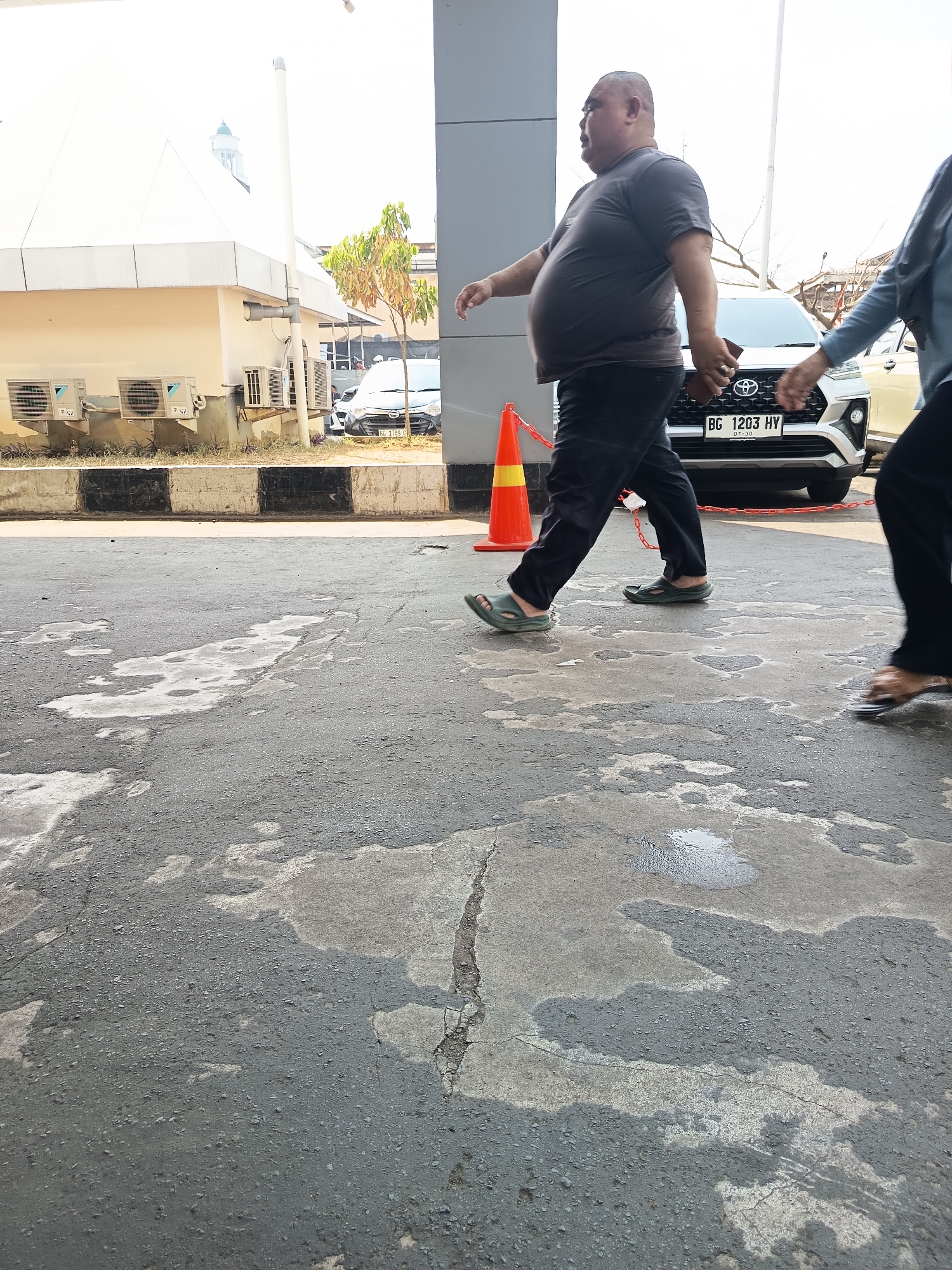 The image shows a man in a grey t-shirt and black pants walking on a wet asphalt surface. He is wearing green sandals. Behind him, a white car with a license plate reading "BG 1203 HY" is parked. To the left of the man, an orange traffic cone is visible. In the background, there is a building with air conditioning units mounted on its exterior wall and a small tree. The scene appears to be a public area, possibly near a building or parking lot. The overall lighting suggests it is daytime. The image is likely taken in Lubuklinggau, Indonesia, as indicated by the license plate format.