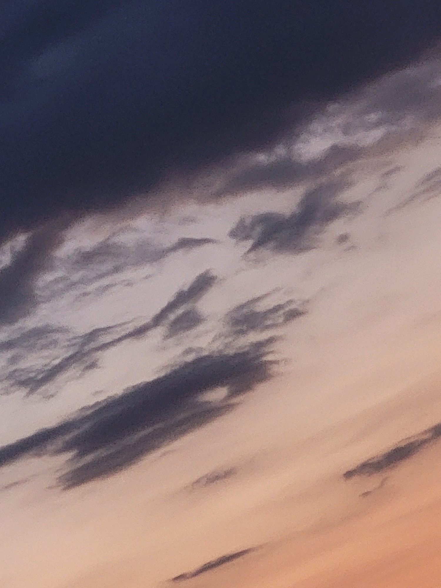 The image is a close-up, abstract view of a sky with clouds. The upper left portion of the image is dominated by dark, almost black, clouds. Moving diagonally downwards and to the right, the clouds become lighter, transitioning through shades of gray and pinkish-orange. There are streaks and wisps of clouds, some thin and feathery, others thicker and more defined, creating a textured and layered effect. The lighting suggests either dawn or dusk. There are no discernible people, objects, or specific landmarks visible, rendering it impossible to determine a location within the city of Ozolnieki, Latvia. The focus is solely on the atmospheric conditions of the sky.