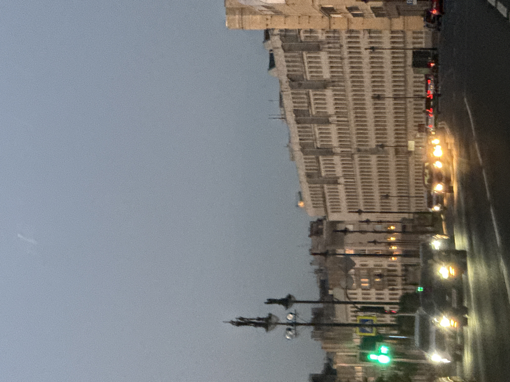 This media file depicts a street scene in Saint Petersburg, Russia, likely during the evening or twilight hours. The dominant subject is a large, multi-story building with a repetitive, horizontal architectural style, possibly a residential complex or an office building. Its facade is pale and appears to be made of concrete or stone. In front of the building, a street is visible, with cars driving and their headlights and taillights illuminated, suggesting low light conditions. There are traffic lights, one of which is green, indicating ongoing traffic flow. Streetlights and possibly lights from within the building also contribute to the illumination of the scene.
The weather appears to be overcast or hazy, given the muted sky color and the lack of distinct shadows. The overall atmosphere is calm and urban, with no discernible human figures or distinct emotional cues. The orientation of the image is vertical, and it is slightly tilted. The perspective is from street level, looking towards the building and down the street. The presence of the building's characteristic architecture, combined with the context of Saint Petersburg, helps to place the scene within this specific Russian city.