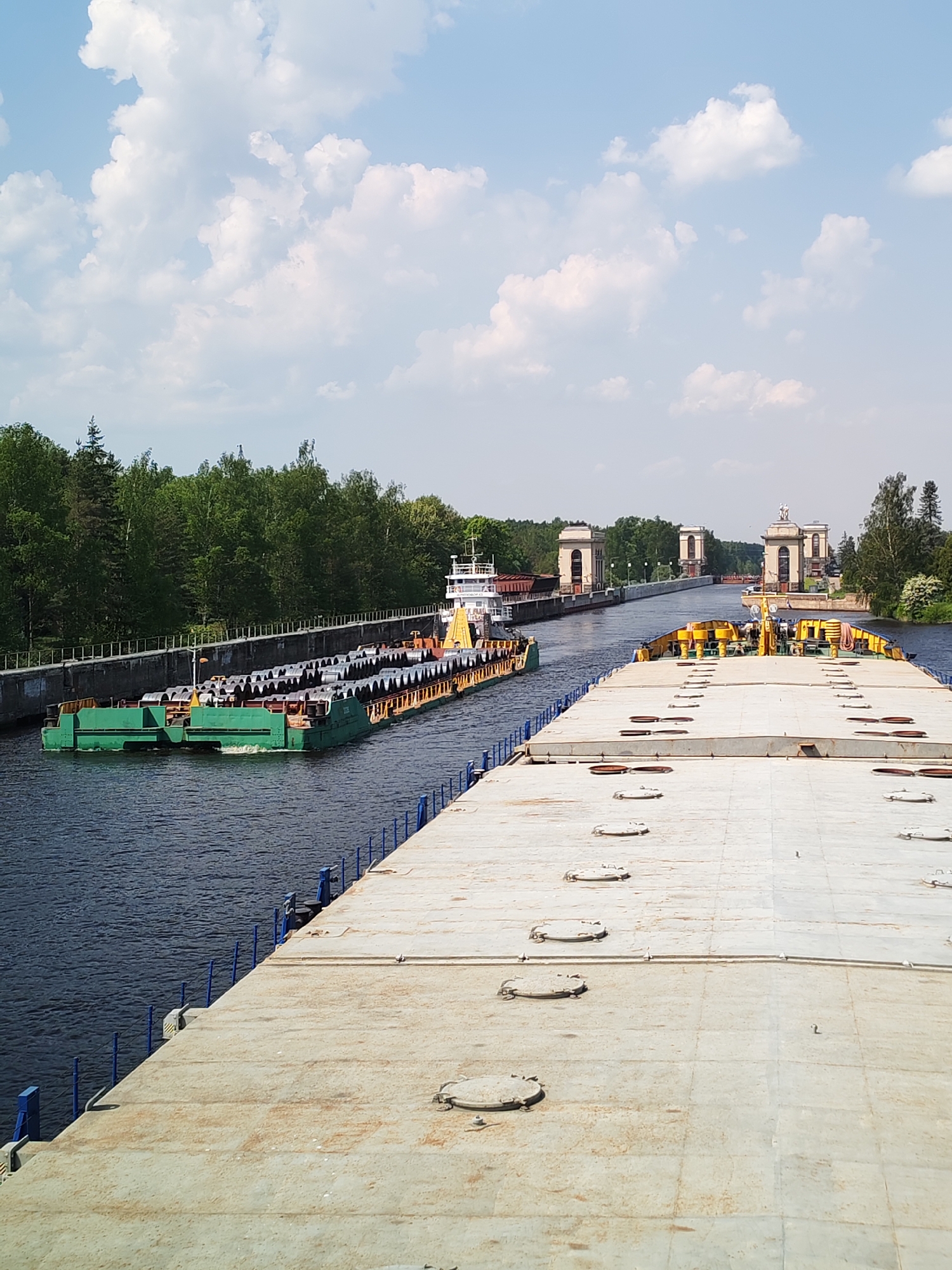 A wide-angle shot shows a waterway with two barges passing through a lock system on a sunny day, presumably in Tempy, Russia. The closest barge, occupying the bottom half of the frame, is a large, flat, concrete-colored vessel. Its deck is segmented into square or rectangular sections, marked with numerous circular access points. The edges are lined with a blue railing system. To the left, a smaller, green barge loaded with what appears to be metal rolls or coils is being pushed or guided by a white and red tugboat. The barges are passing through a canal lined with concrete walls and lush green trees. In the distance, the canal stretches towards three sets of matching ornate lock structures, each featuring arched openings and decorative elements at the top. The sky above is blue with scattered puffy white clouds, indicating a clear day.