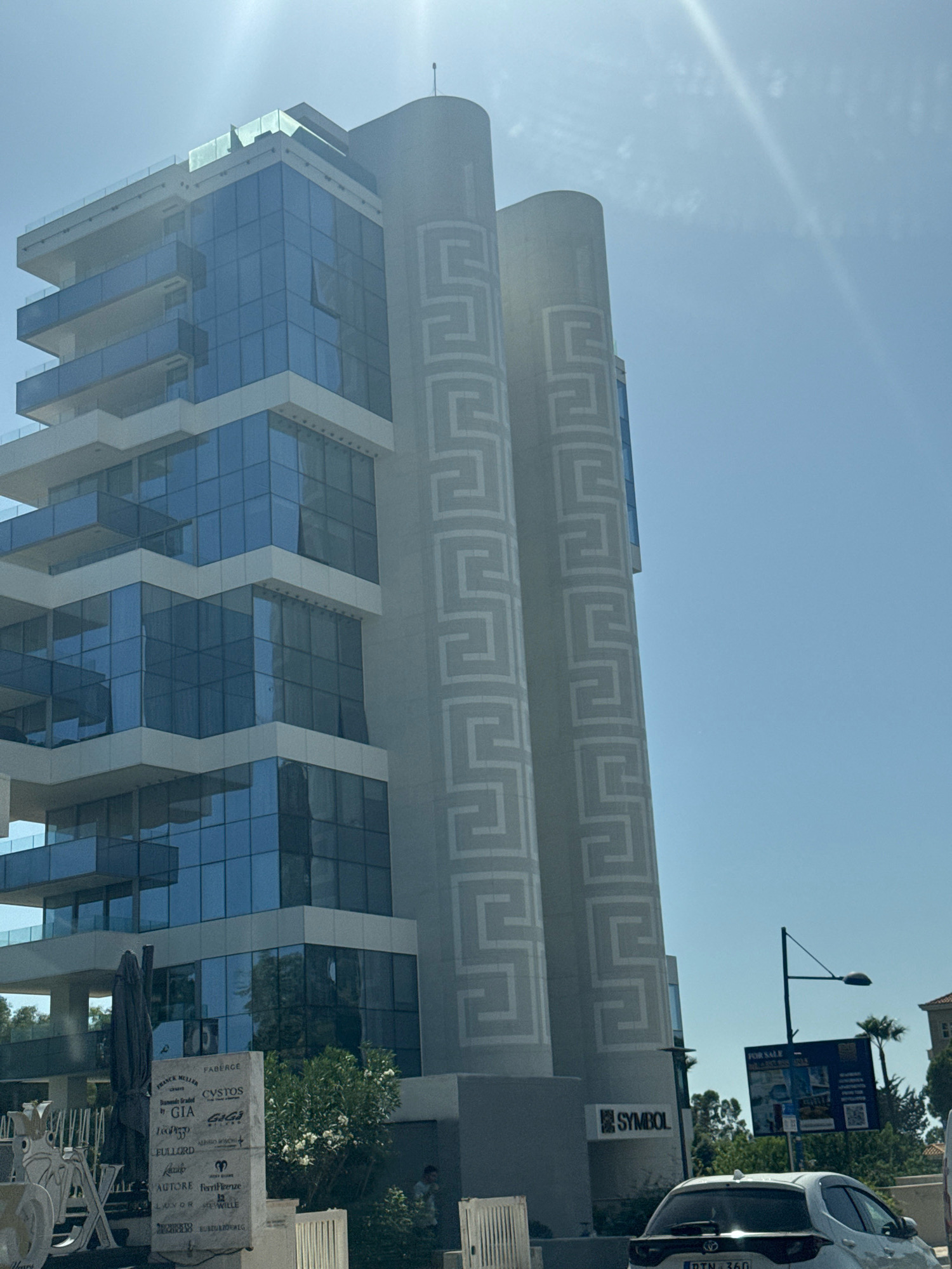 An eye-level, medium shot captures a modern, multi-story building with a glass facade and a decorative cylindrical element featuring a repeating geometric pattern. The building is situated in a sunlit urban environment, with a clear blue sky dominating the upper portion of the frame. A white car is parked in the foreground. The location appears to be Agios Tychona Community, Cyprus, as indicated by signs visible on a nearby stone monument and a "FOR SALE" billboard. The overall scene suggests a commercial or residential area, possibly near the coast due to the presence of palm trees.